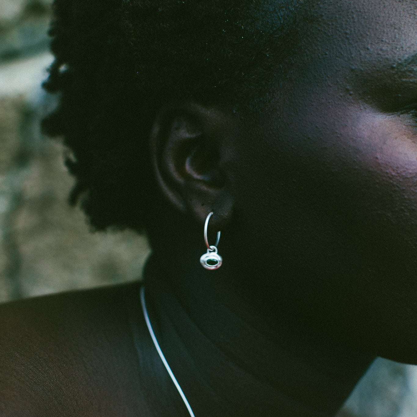 A close-up side profile of a person wearing a small silver hoop earring with a dangling round silver charm, against a dark, moody background.