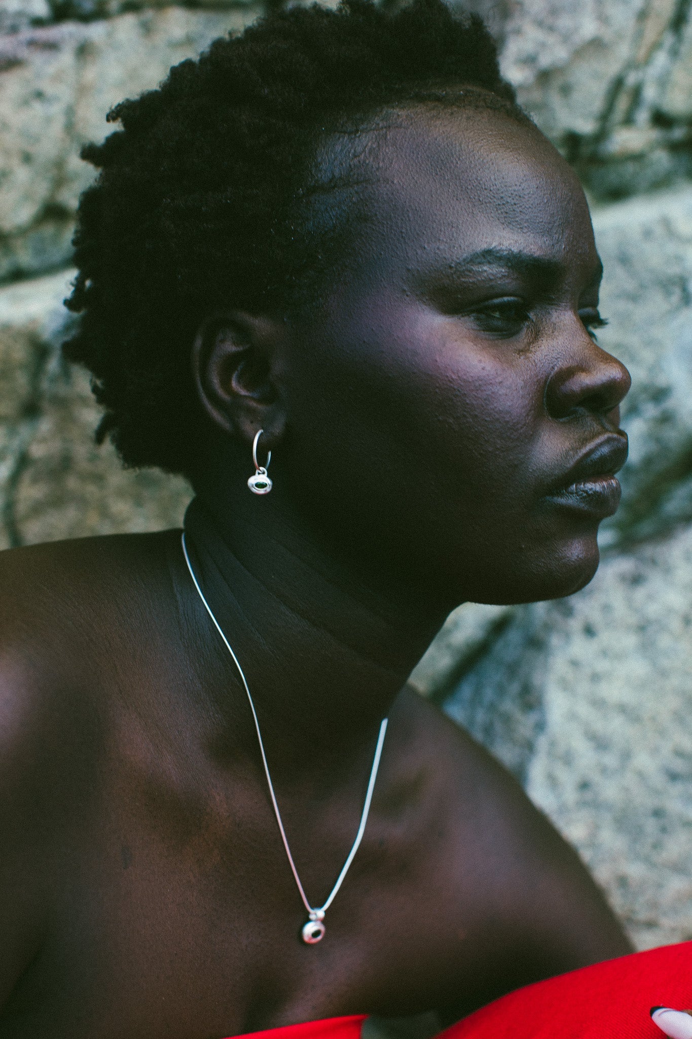 A close-up side profile of a woman with short dark curly hair, wearing a delicate silver necklace with a small red and white gemstone pendant and a matching silver hoop earring, set against a textured gray stone wall.