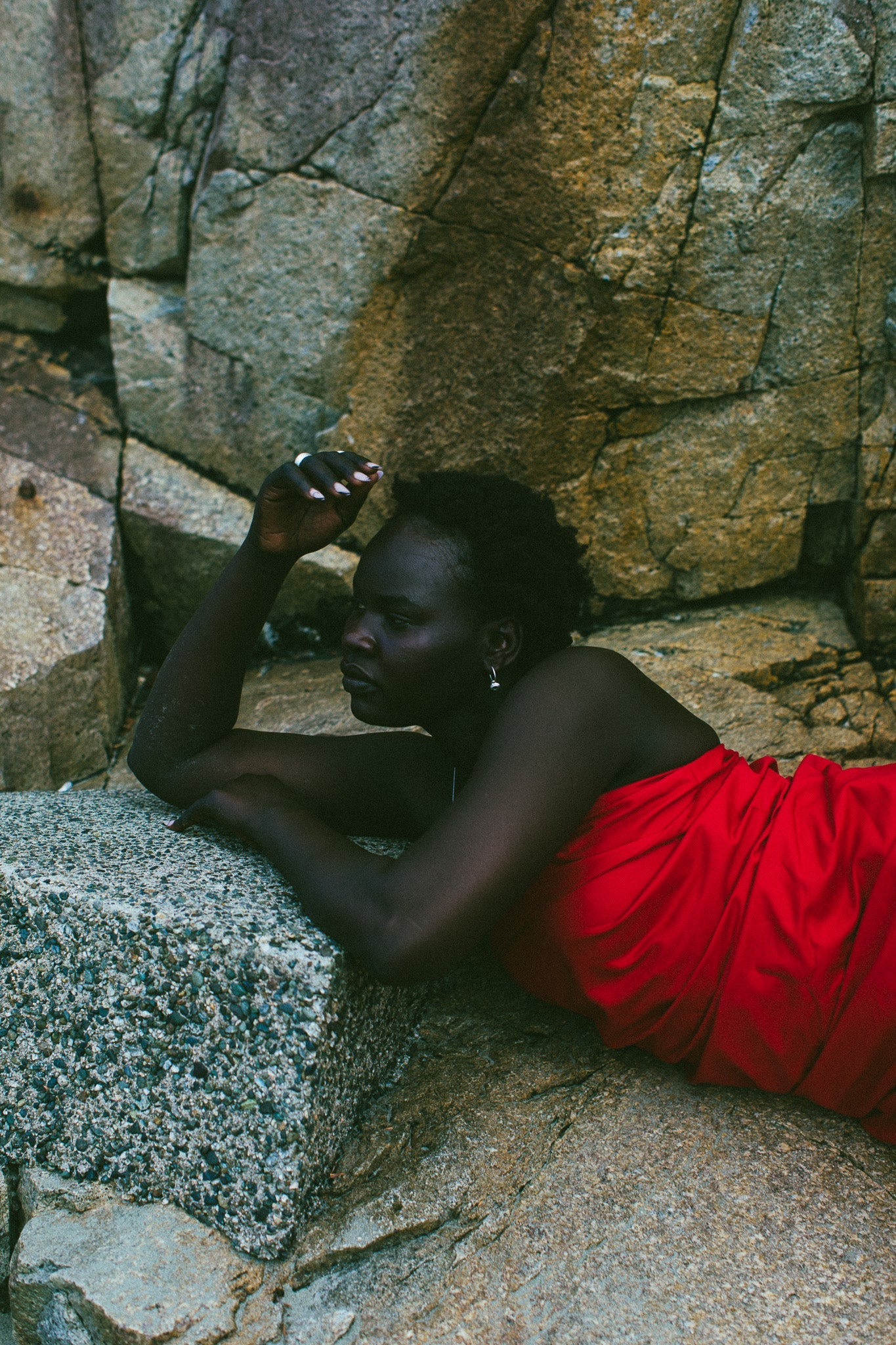 A woman reclining on large stone blocks outdoors, wearing a bright red draped garment. The background consists of natural rock formations.