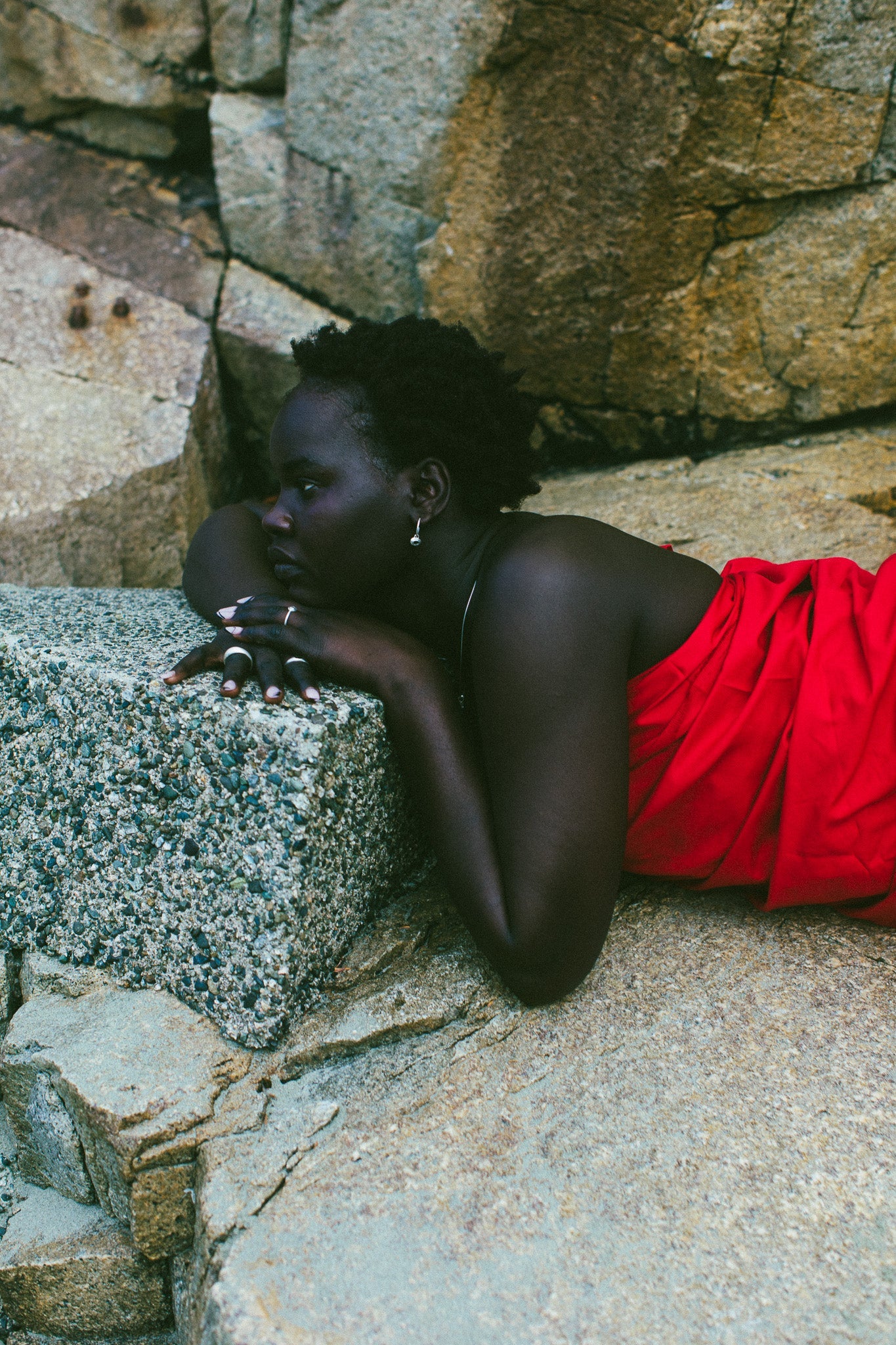 A woman with short dark hair lies on large gray rocks, wearing a bright red draped garment and small hoop earrings.