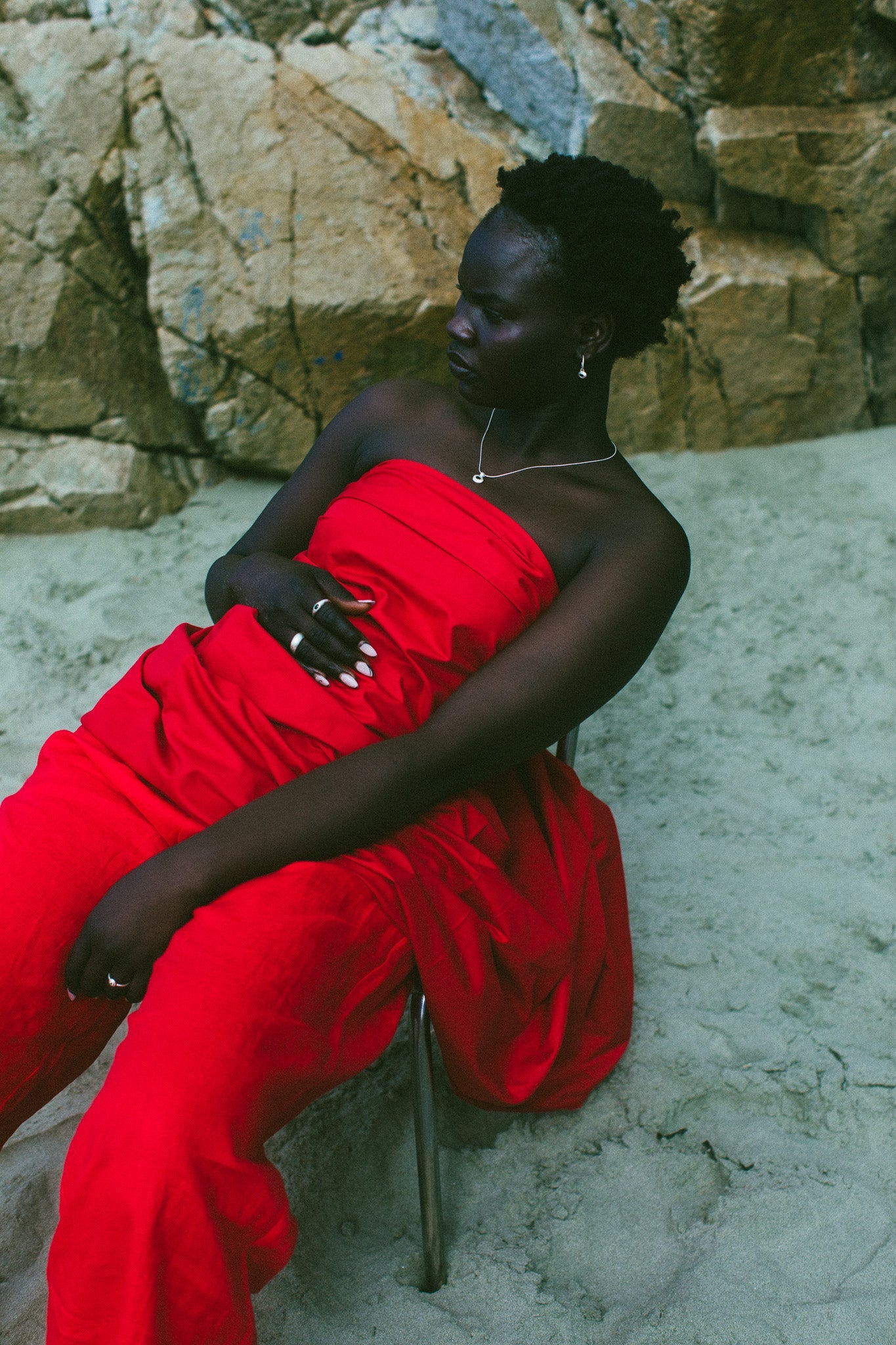 A woman seated outdoors on a metal chair against a rocky, sandy background, wearing a bright red strapless draped dress.