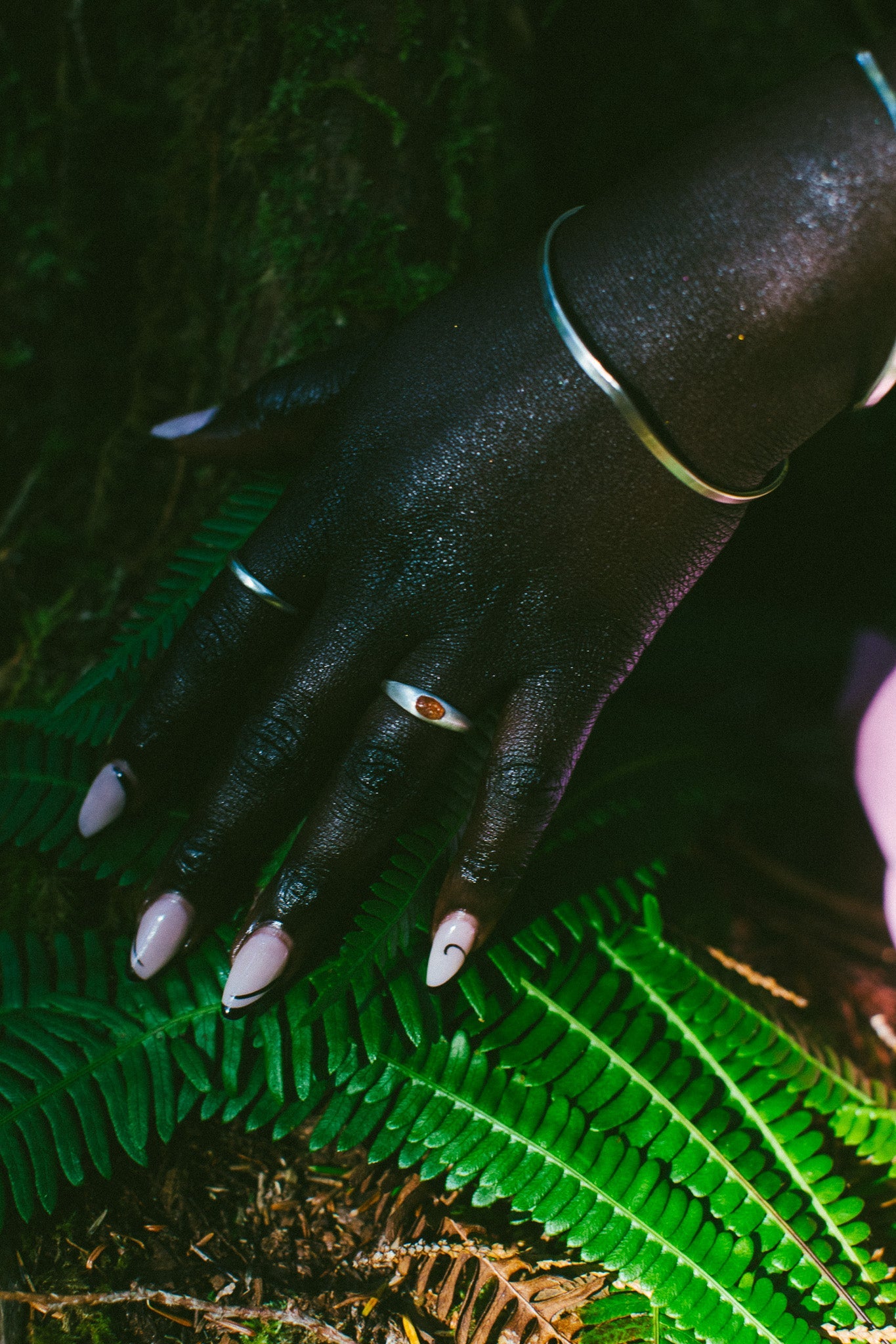 A close-up of a hand resting on bright green fern leaves outdoors. The hand is adorned with silver-toned jewelry, including a bangle bracelet and rings. One ring with an orange oval gemstone.