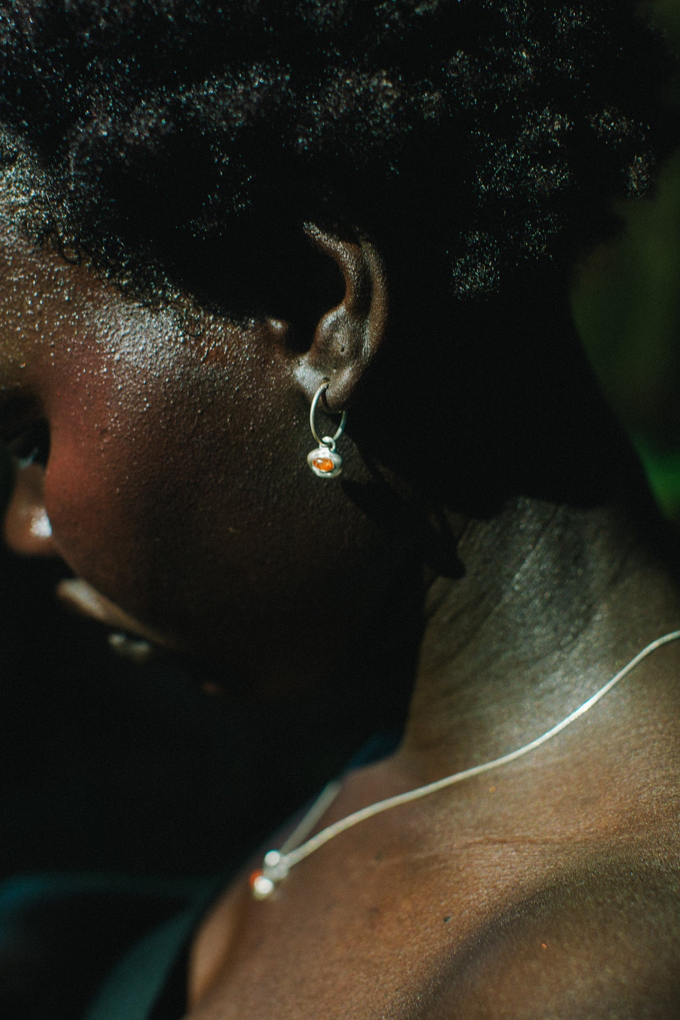 Close-up side profile of a person wearing a small silver hoop earring with a tiny orange and white stone, along with a thin silver necklace.