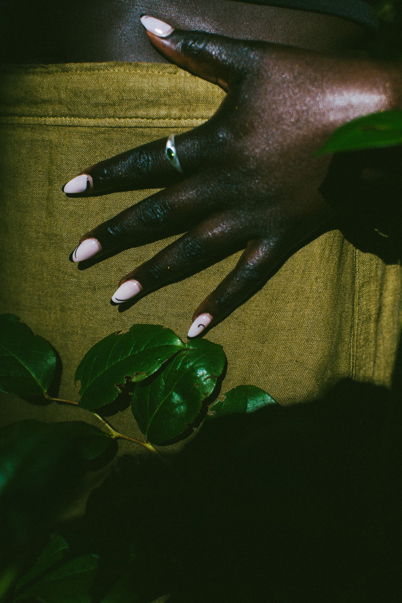 A close-up of a hand with long, pale pink manicured nails resting on a textured olive green fabric surface, with dark green leaves in the foreground and a silver ring on one finger.