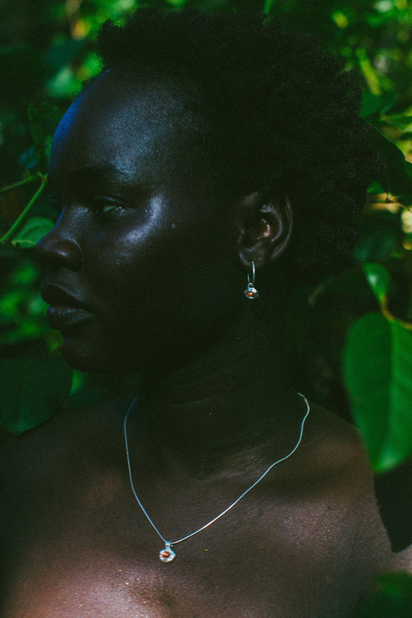 A close-up side profile of a person outdoors among green leaves, wearing a delicate silver necklace with a small round pendant and a matching silver hoop earring.