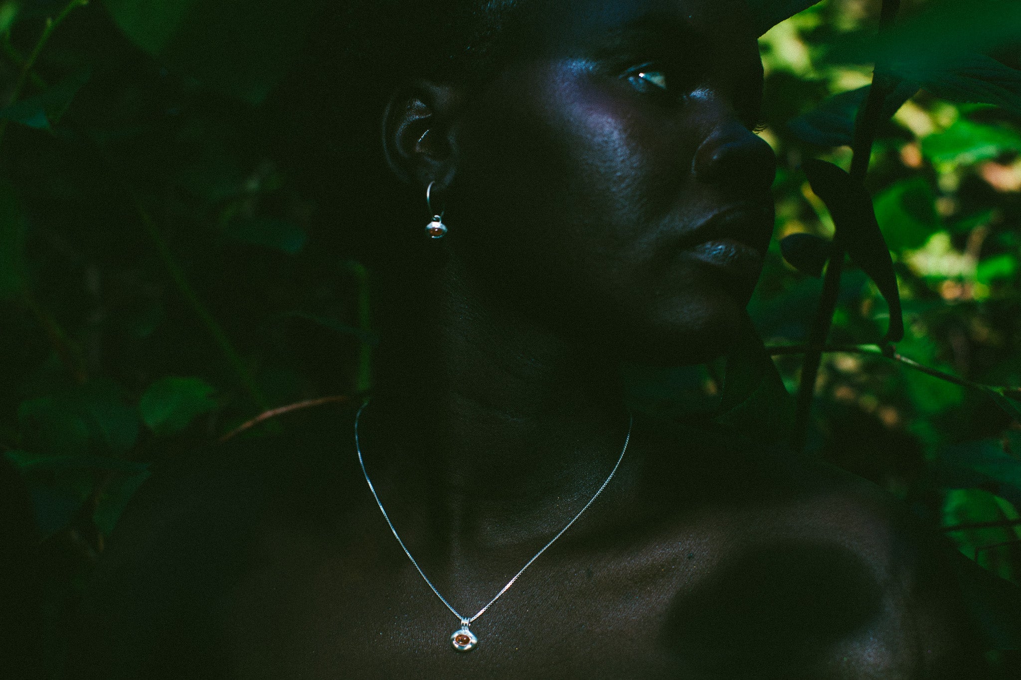 A close-up portrait of a woman in profile wearing a delicate silver pendant necklace and matching drop earring, set against a dark, leafy outdoor background.