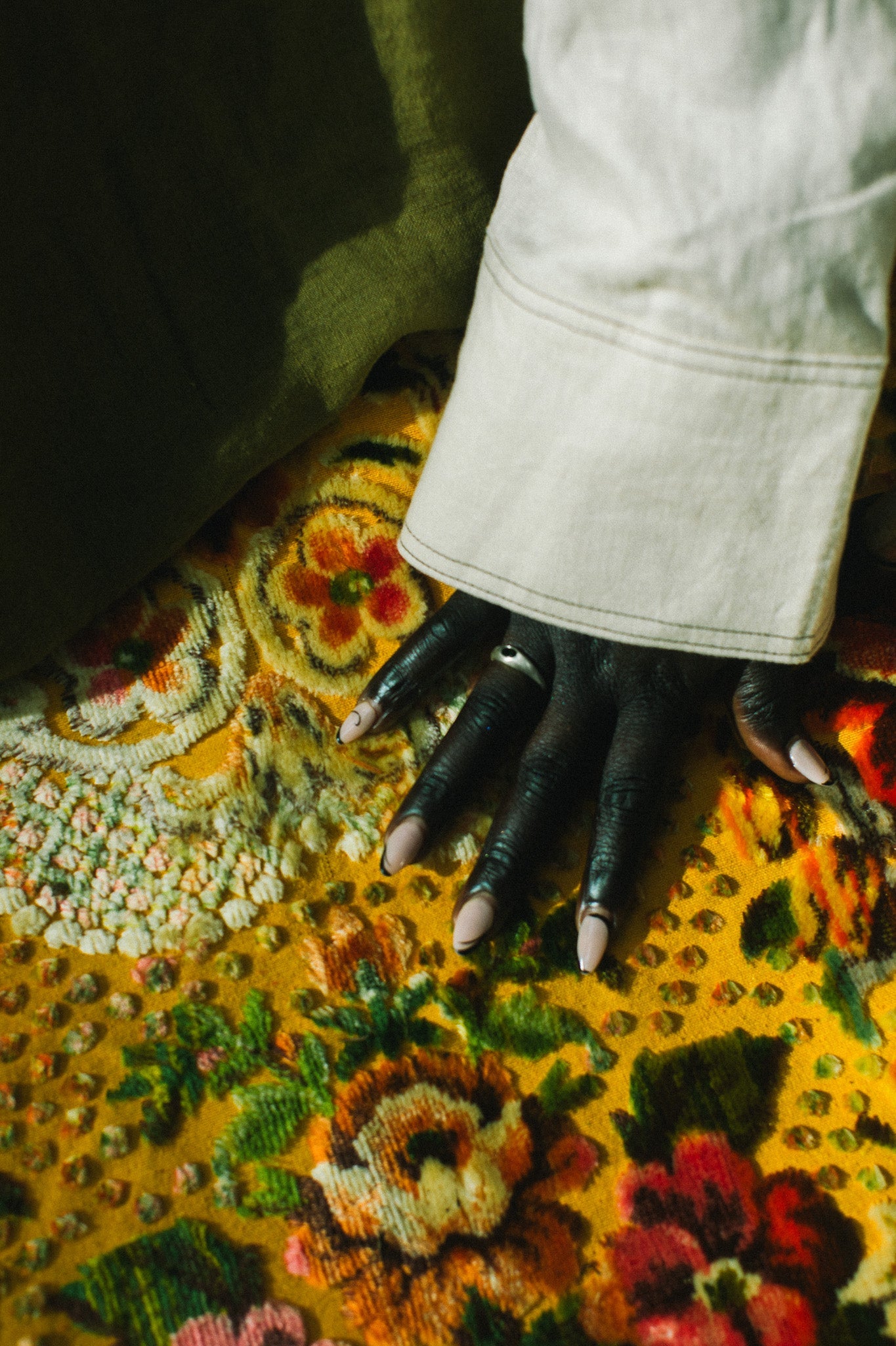 A close-up of a hand wearing a silver ring with an oval, green gemstone resting on a vibrant, multicolored floral patterned rug, with a light-colored denim cuff visible at the wrist.