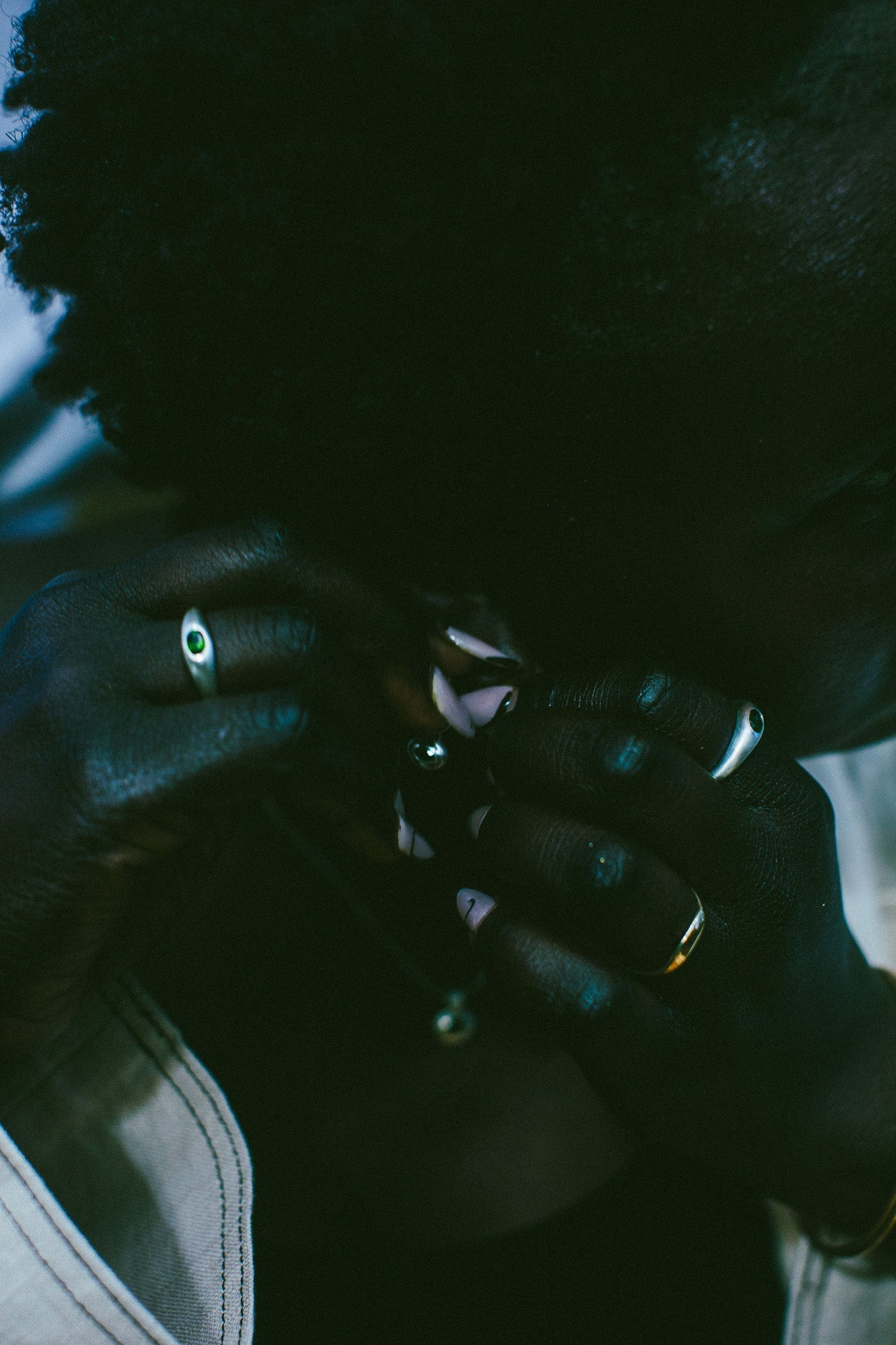 A close-up lifestyle shot of a person wearing multiple rings, including a prominent green stone ring on one finger, in low, moody, dark teal-toned lighting.