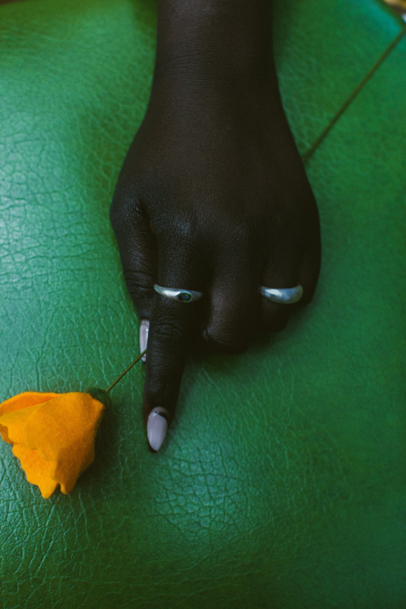 A close-up of a hand resting on a textured green surface, wearing two silver rings, with a small yellow flower nearby.