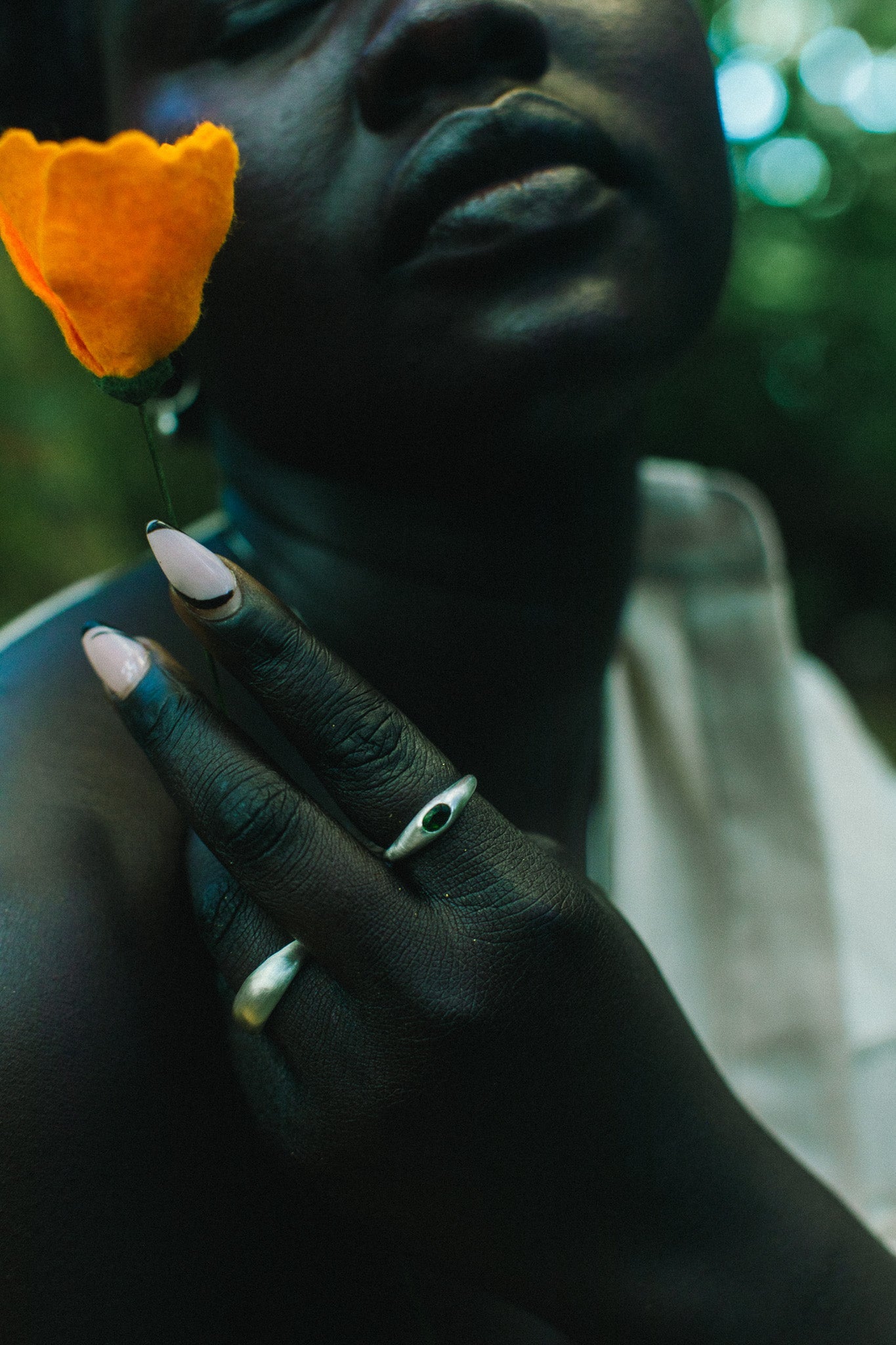 A close-up lifestyle shot of a person wearing two silver rings on their fingers, holding a bright orange flower near their face. The background is a dark, blurred outdoor setting with green foliage.