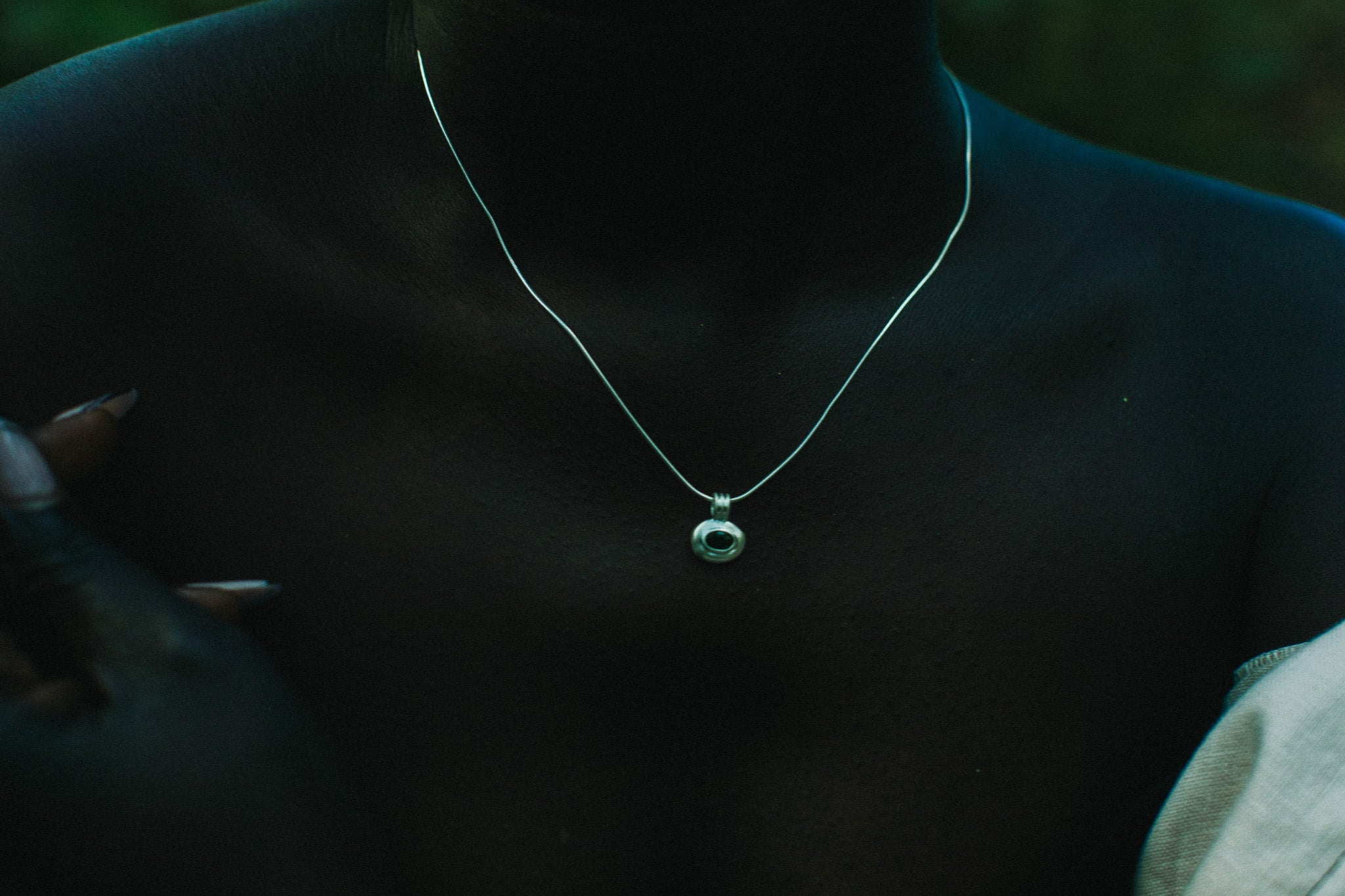 A close-up of a person wearing a delicate silver chain necklace with a small circular pendant featuring a green stone.
