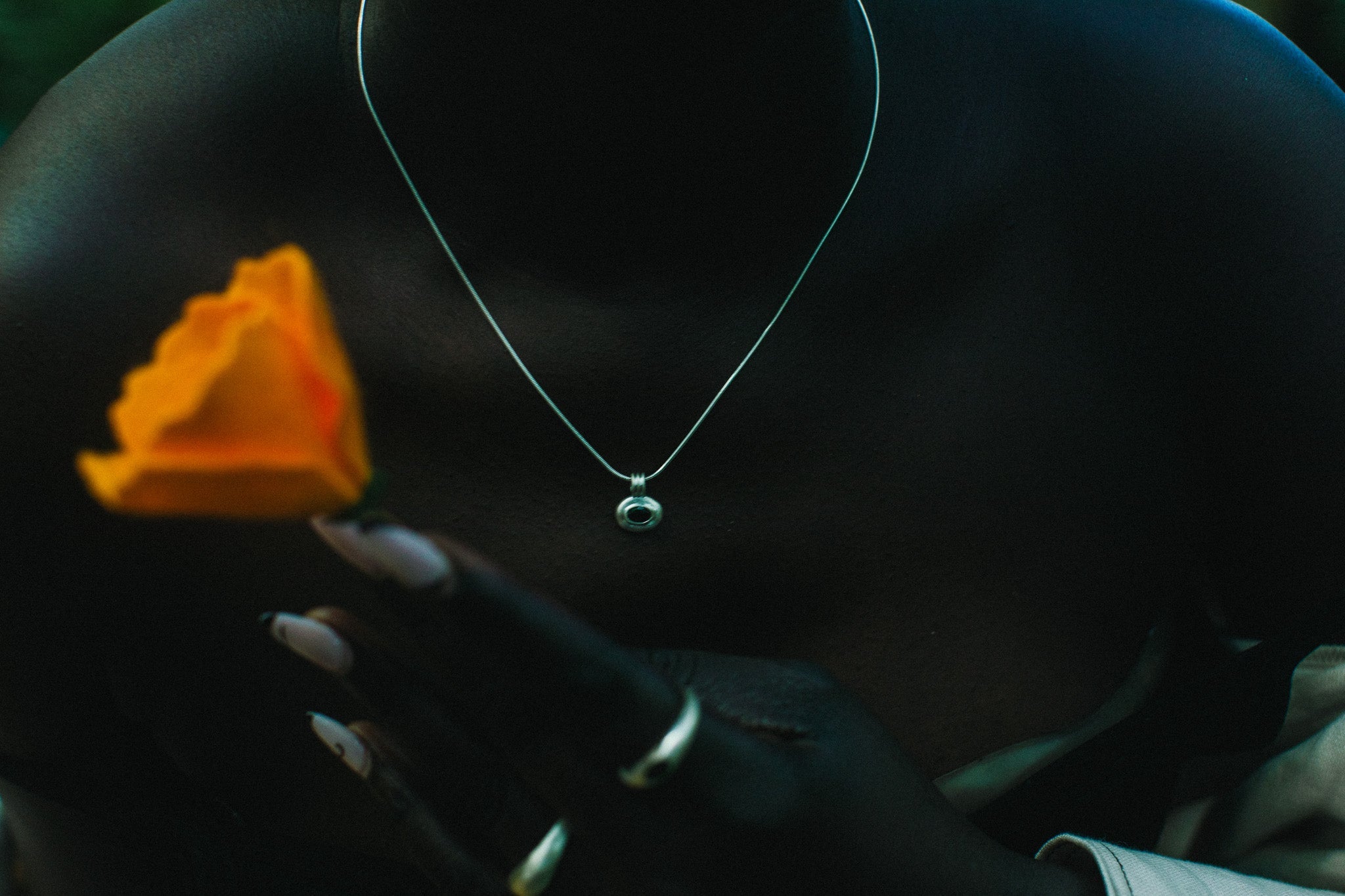 A close-up lifestyle shot of a person wearing a delicate silver chain necklace with a small round pendant, holding a vibrant orange rose. 