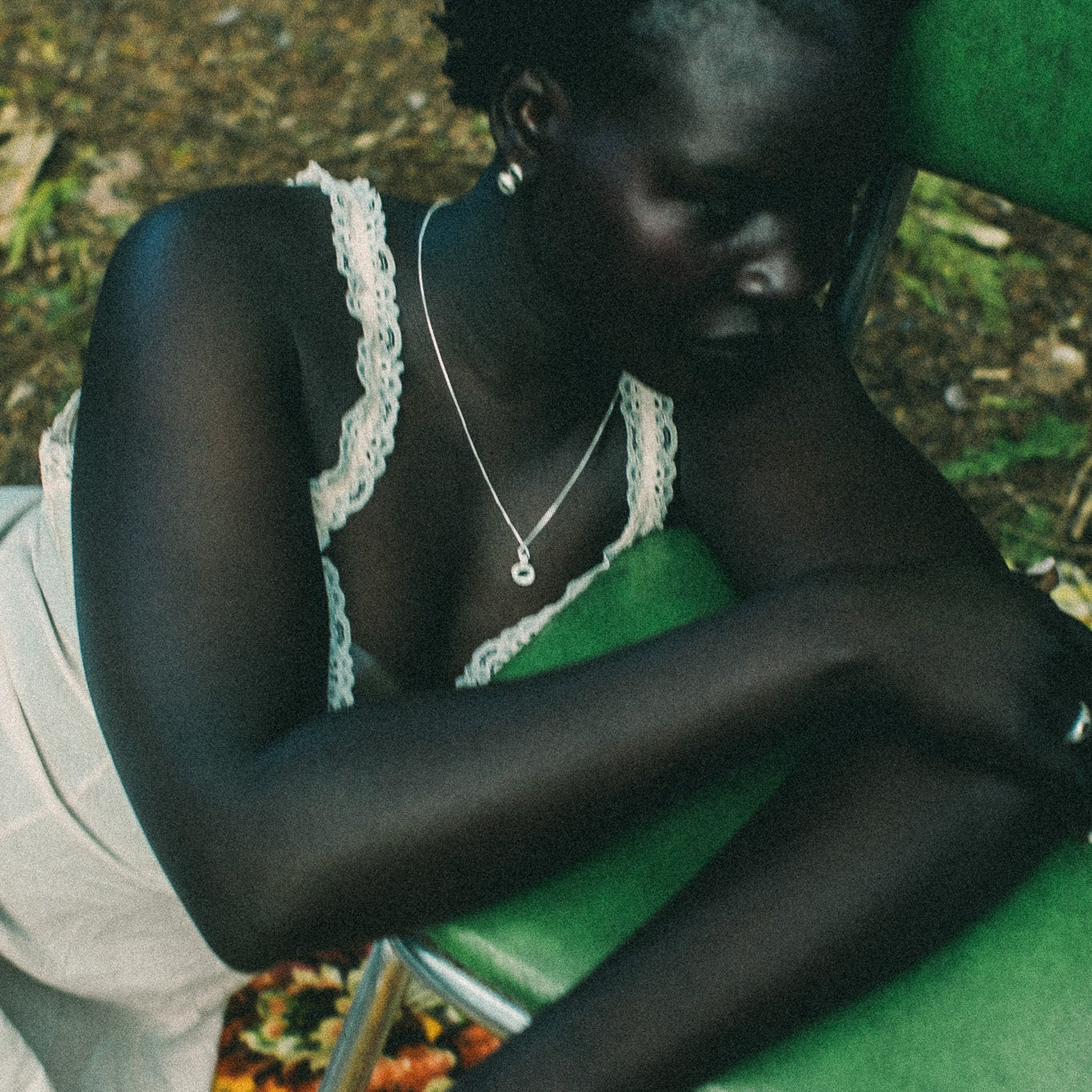 A woman rests her head on a green chair in an outdoor setting, wearing a white lace-trimmed camisole and a silver necklace, and a ring.