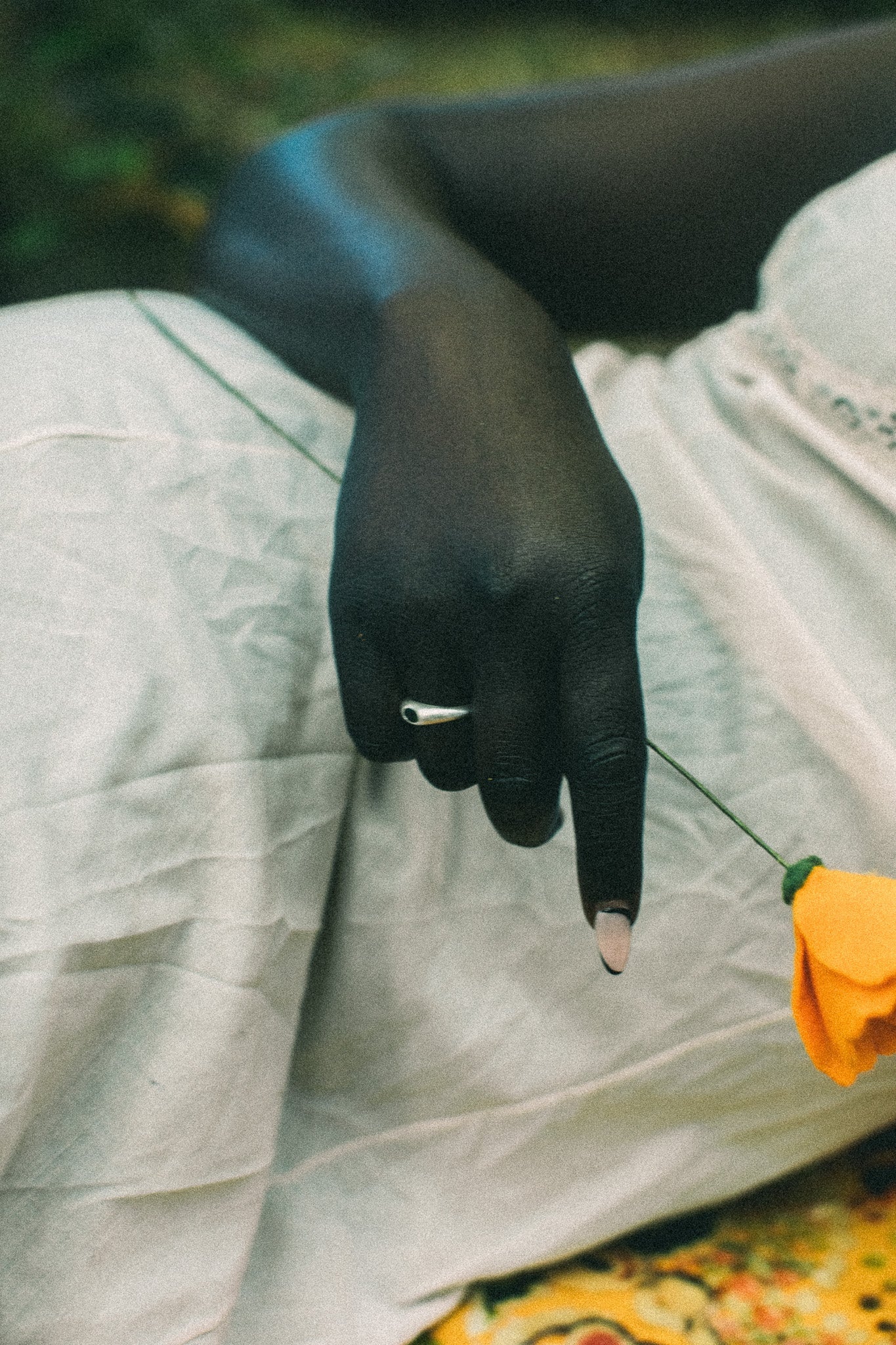 A close-up lifestyle shot of a person's hand resting on their lap, wearing a silver ring, with a small yellow flower held near their finger. They are dressed in a white skirt, set against a soft-focus outdoor background.