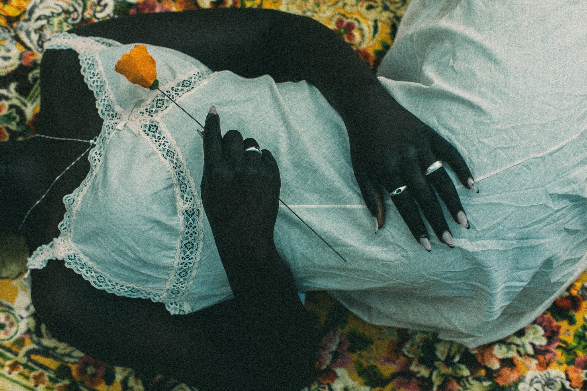 A person lying on a floral blanket, wearing a white lace-trimmed top and light gray skirt, with a single orange flower held near their chest. they are wearing rings.