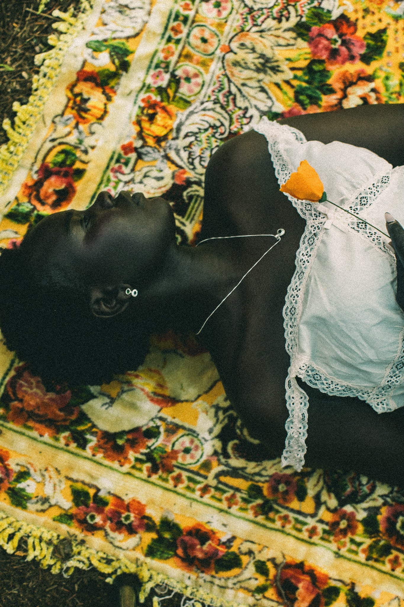A person lying on their back on a colorful floral patterned blanket, wearing a white lace-trimmed top and a small orange flower tucked into the neckline.