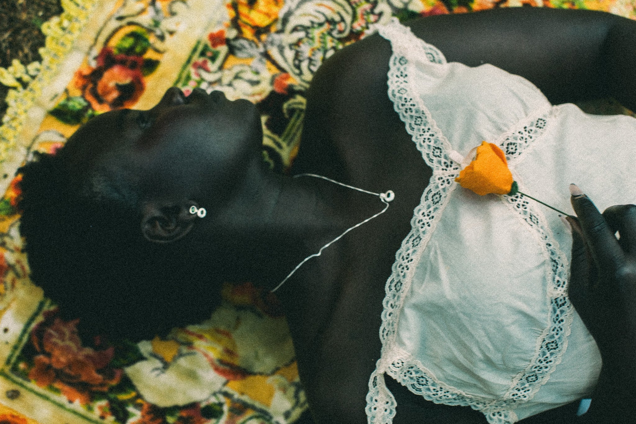 A close-up lifestyle shot of a person lying down wearing a white lace-trimmed top, with a small orange flower pinned to the chest, against a colorful patterned textile background.