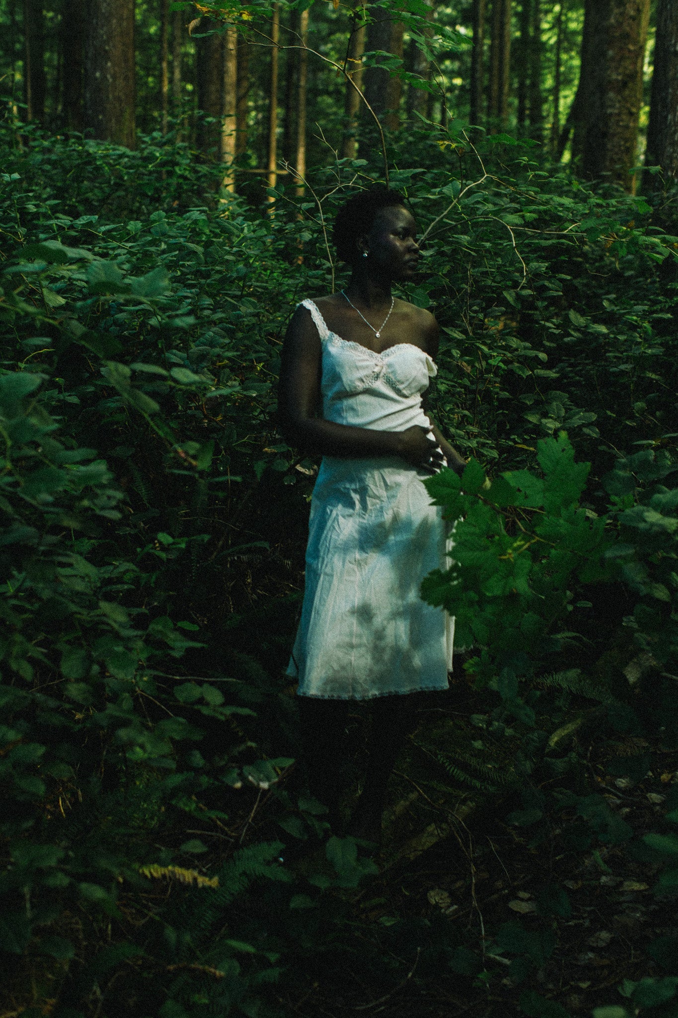 A woman stands in a dense green forest wearing a silver necklace and a white sleeveless dress, surrounded by lush foliage and trees.