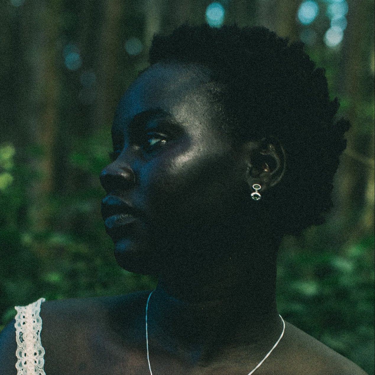 A woman in a white lace-trim top wearing a delicate silver pendant necklace and matching silver stud dangle earrings, photographed outdoors in a dark, green-toned forest setting.