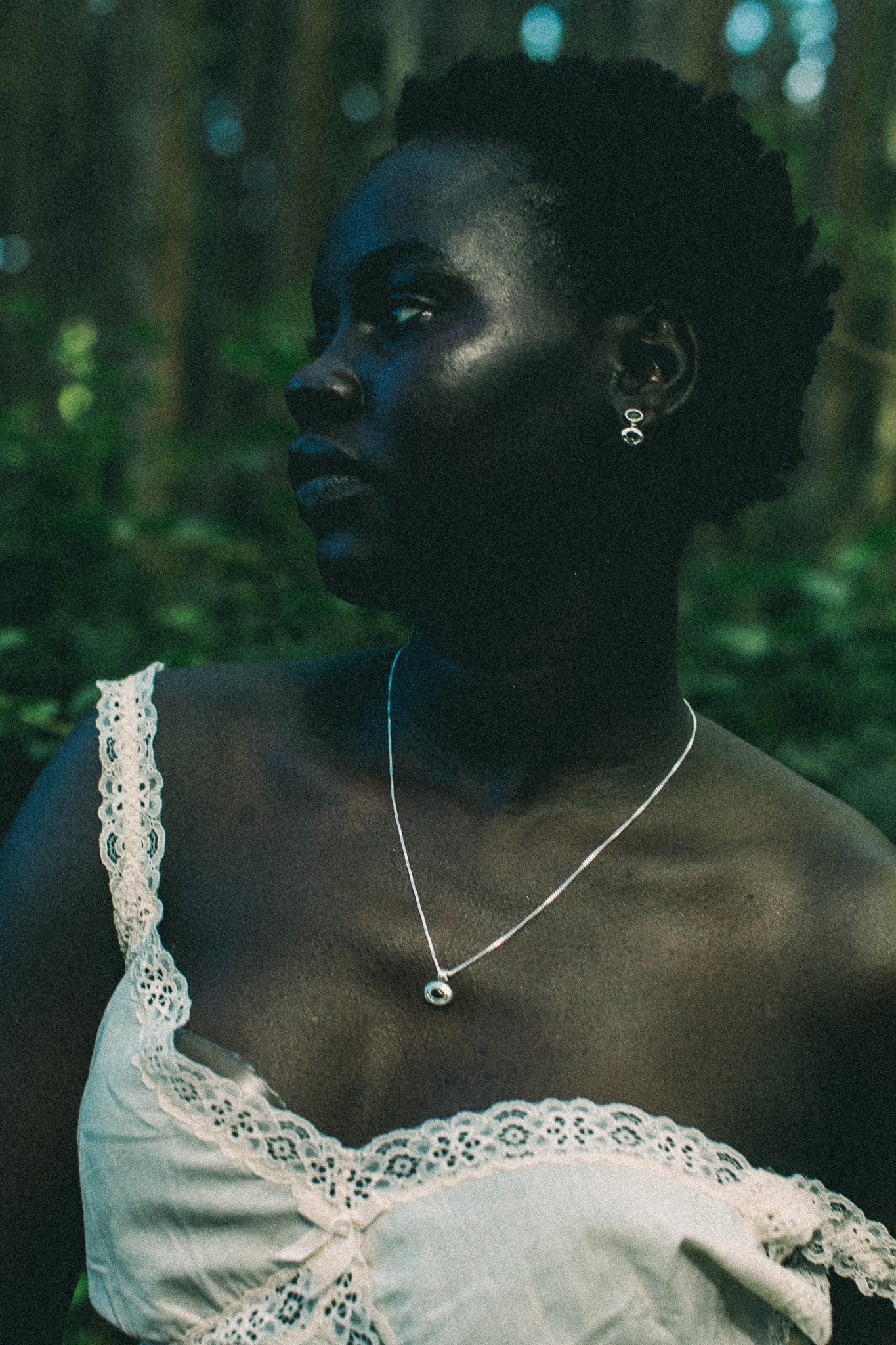 A woman in a white lace-trim top wearing a delicate silver pendant necklace and matching silver hoop earrings, photographed outdoors in a dark, green-toned forest setting.