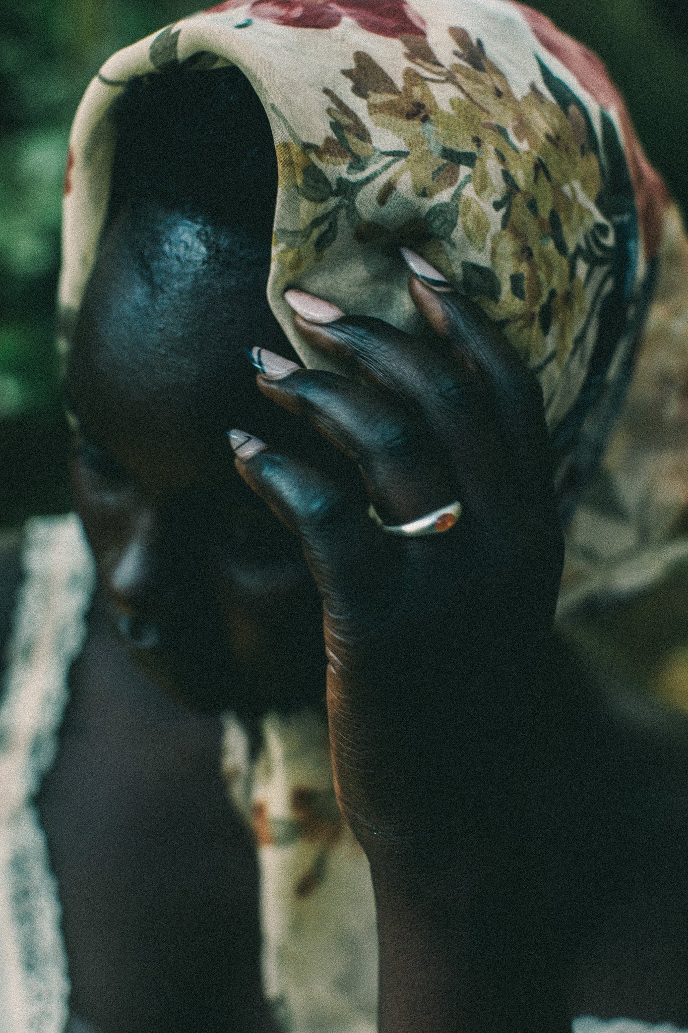 A close-up portrait of a person wearing a patterned headscarf in earthy tones and a silver ring with a small orange stone, with their hand resting against their face.