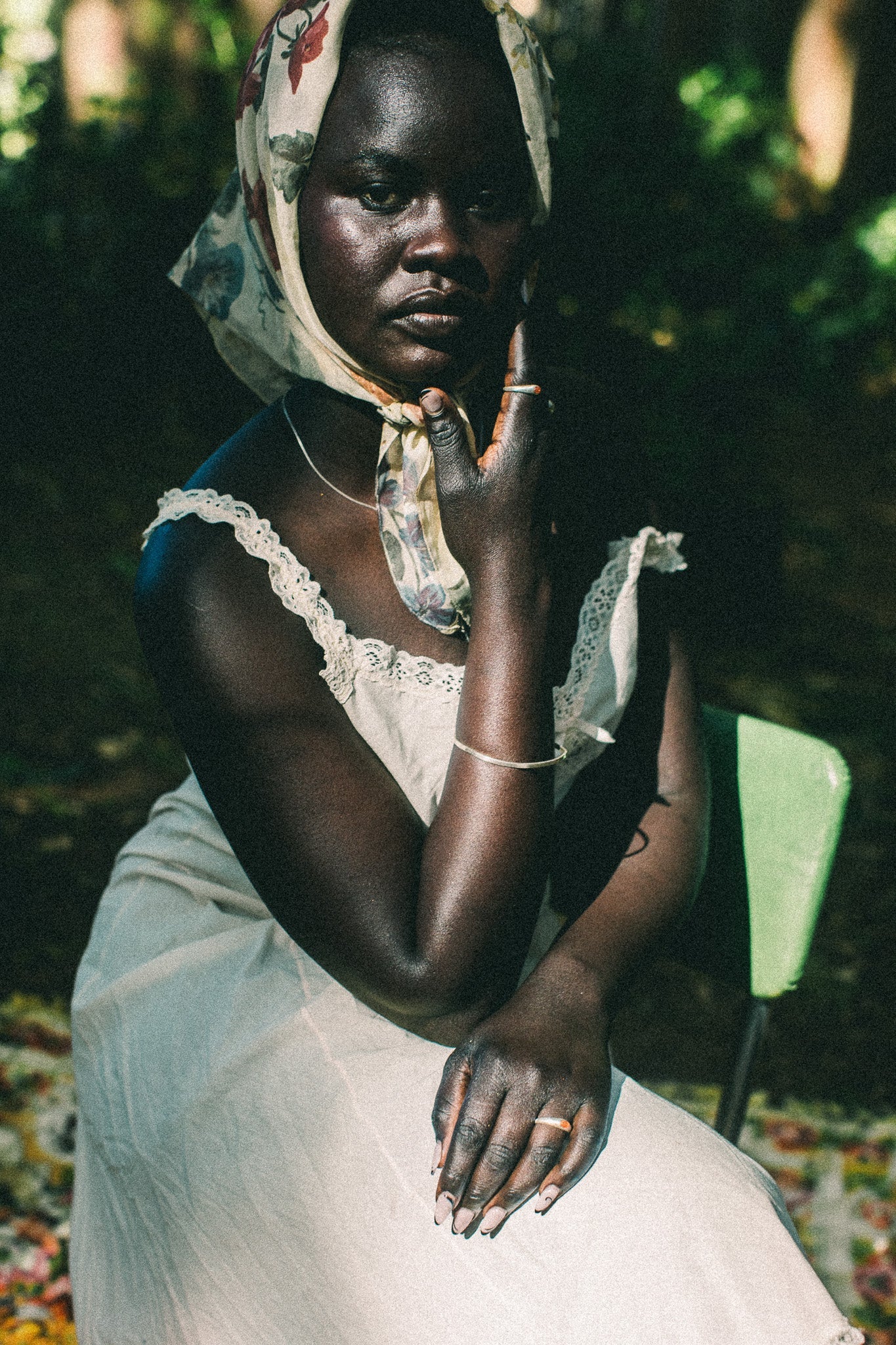 A woman seated outdoors wearing a cream lace-trimmed dress and a patterned headscarf, with dark foliage and a green chair in the background.