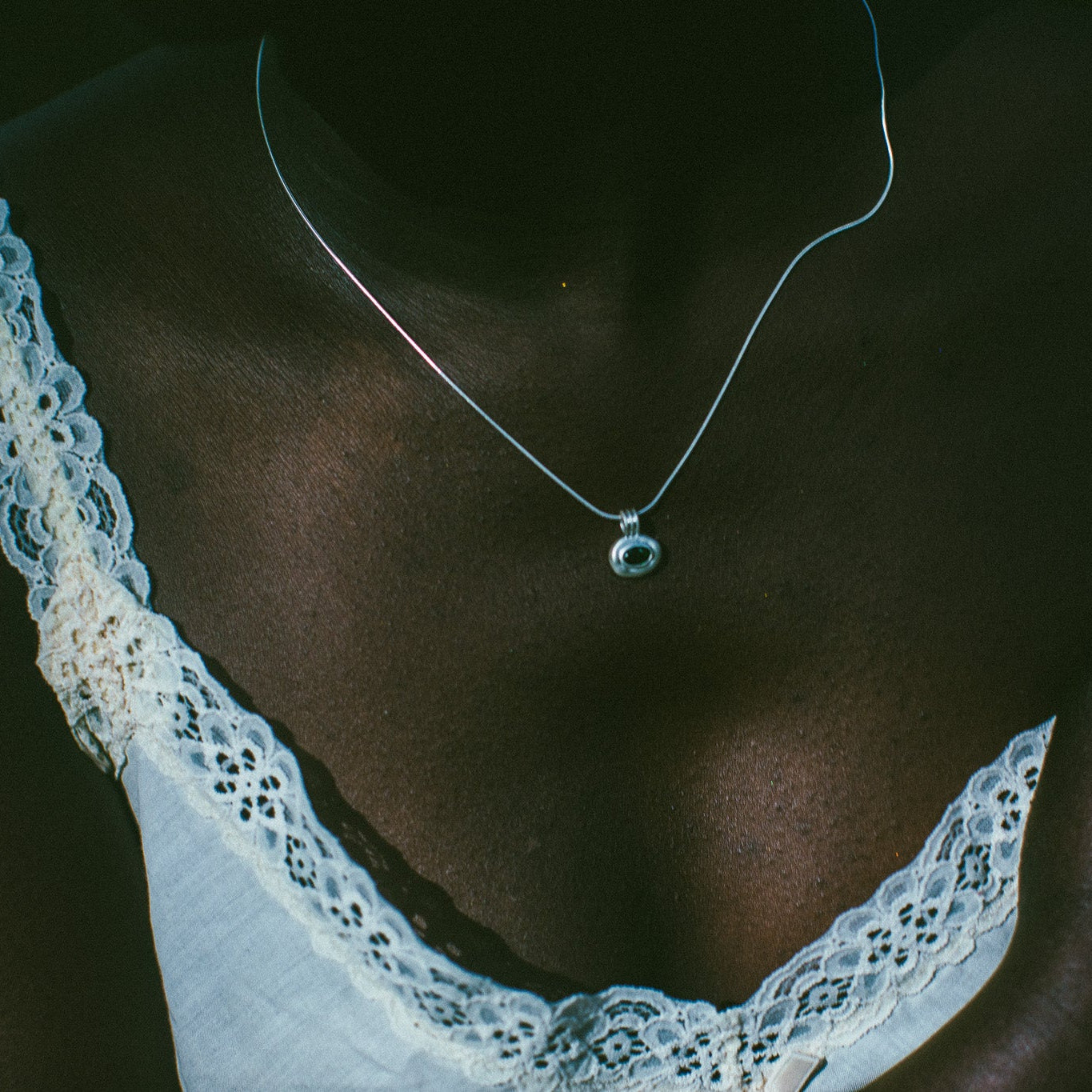 A close-up of a person's chest wearing a delicate silver chain necklace with a small circular pendant, paired with a white lace-trimmed top 