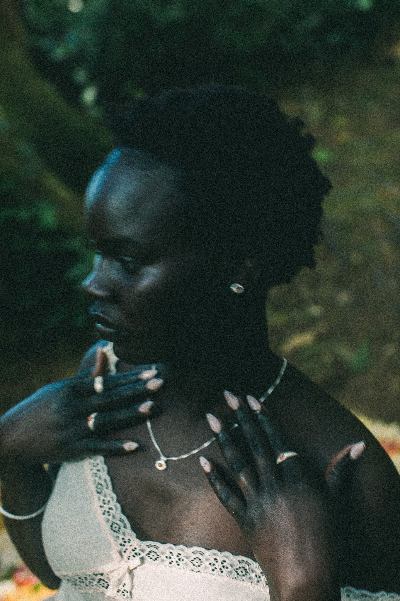 A woman in profile wearing a delicate silver necklace and rings, with her hands near her chest, set against a dark, blurred outdoor background.