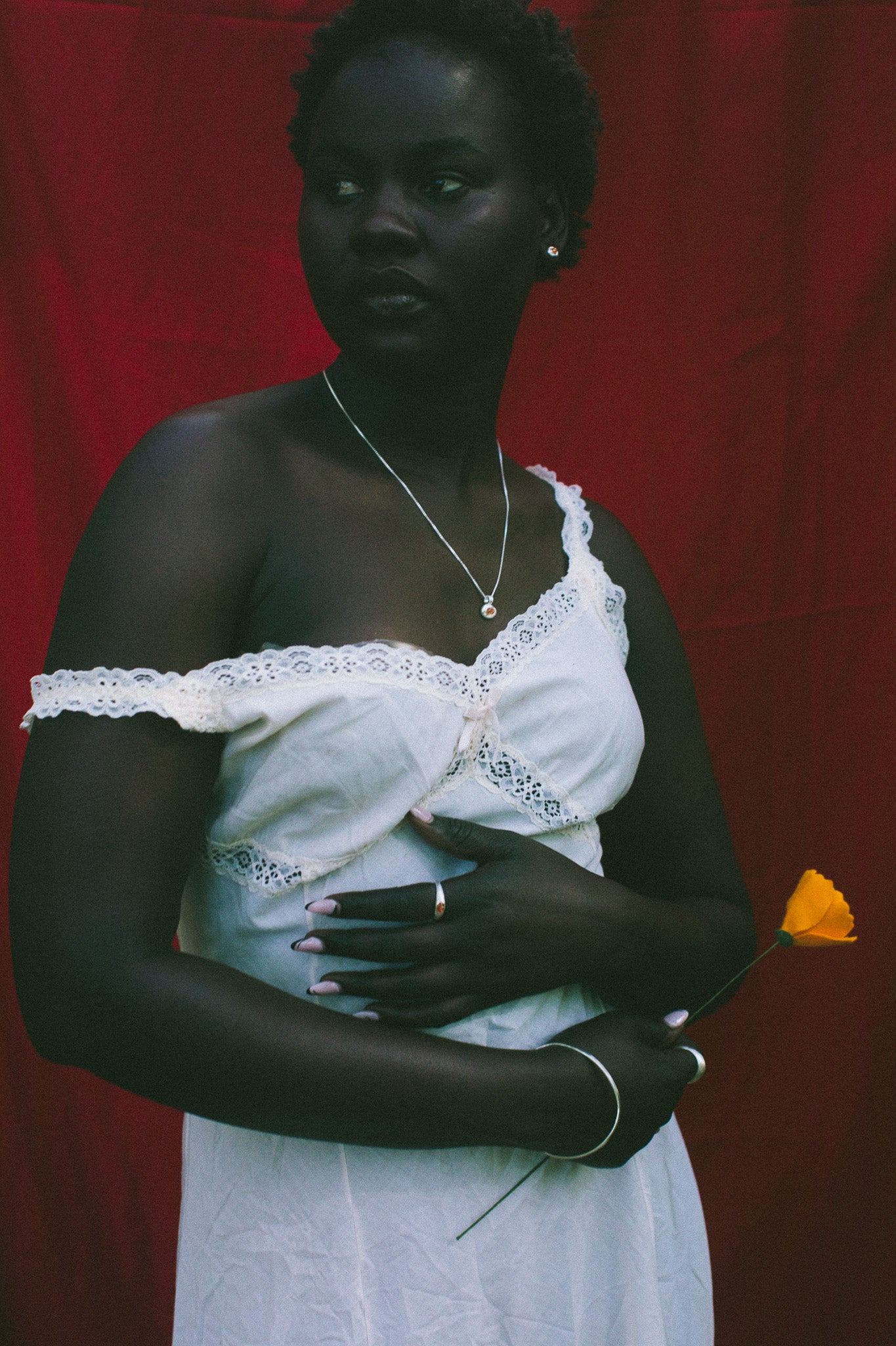 A woman stands against a red fabric backdrop wearing a white one-shoulder dress with lace trim and holding a small yellow flower.