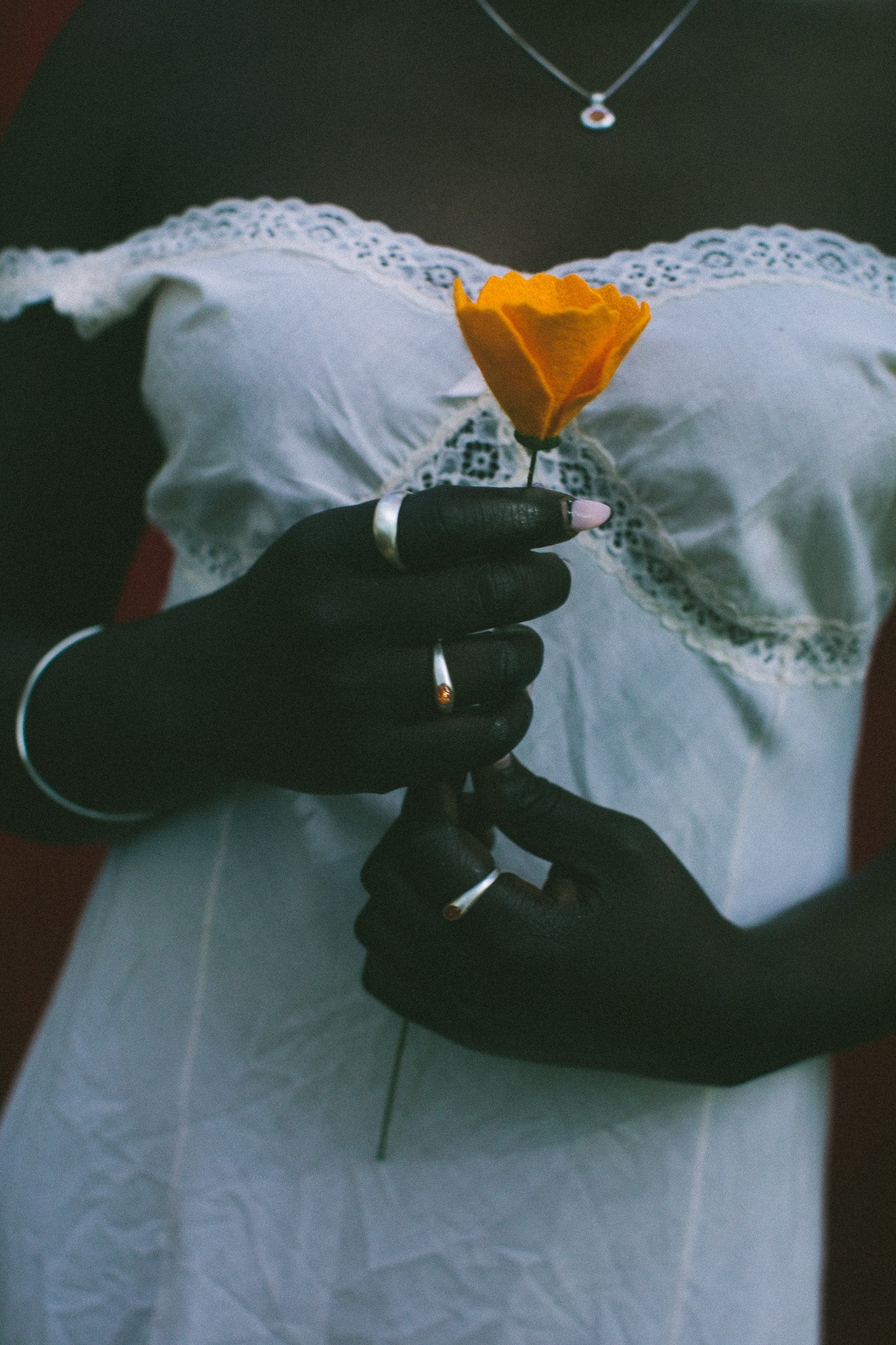 A close-up of a person wearing a white lace-trimmed dress, holding a single orange flower. The person is also wearing several silver rings and a delicate necklace.