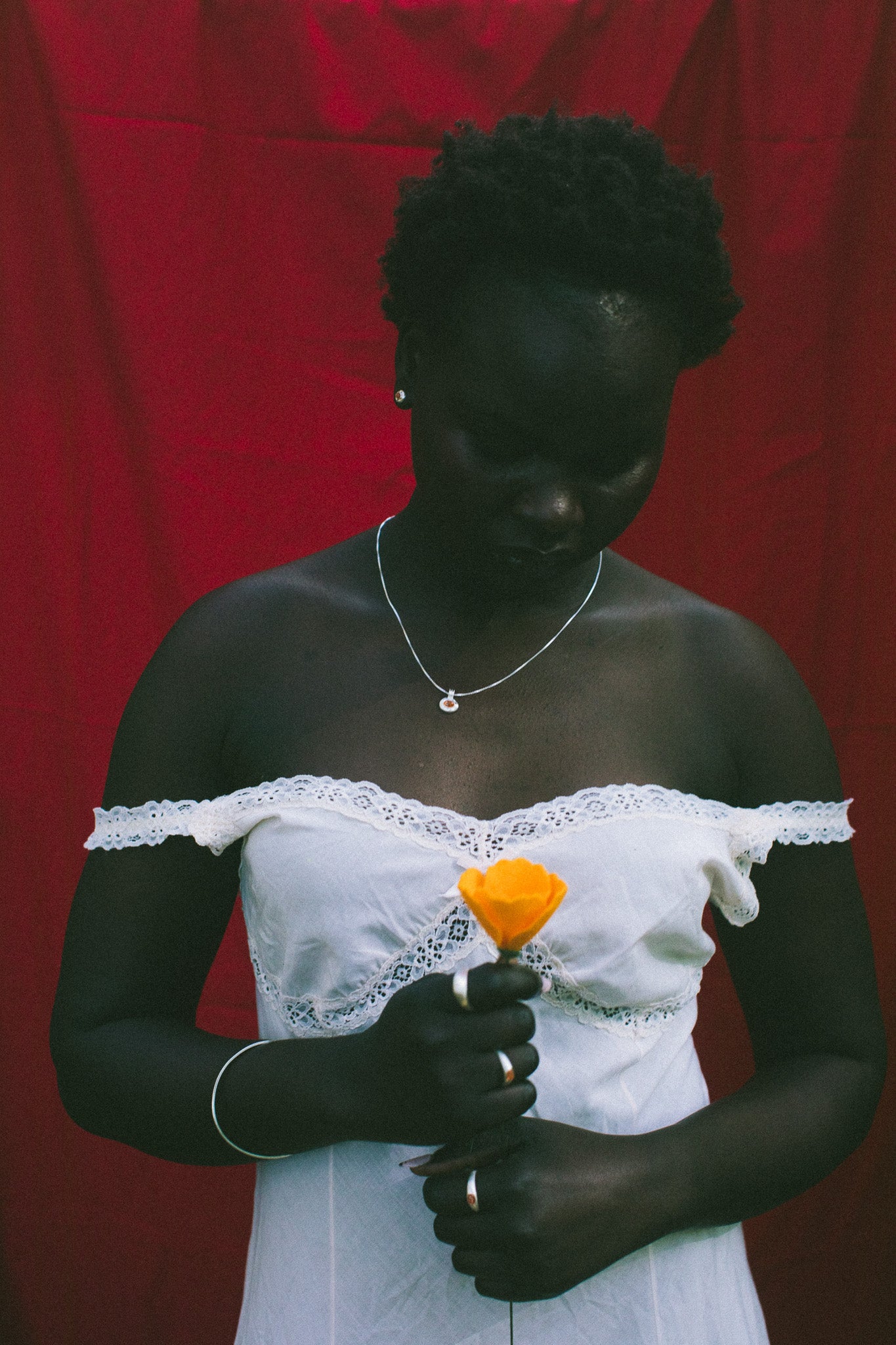 A woman wearing a white off-the-shoulder lace dress, holding a bright orange flower in front of a red draped backdrop.
