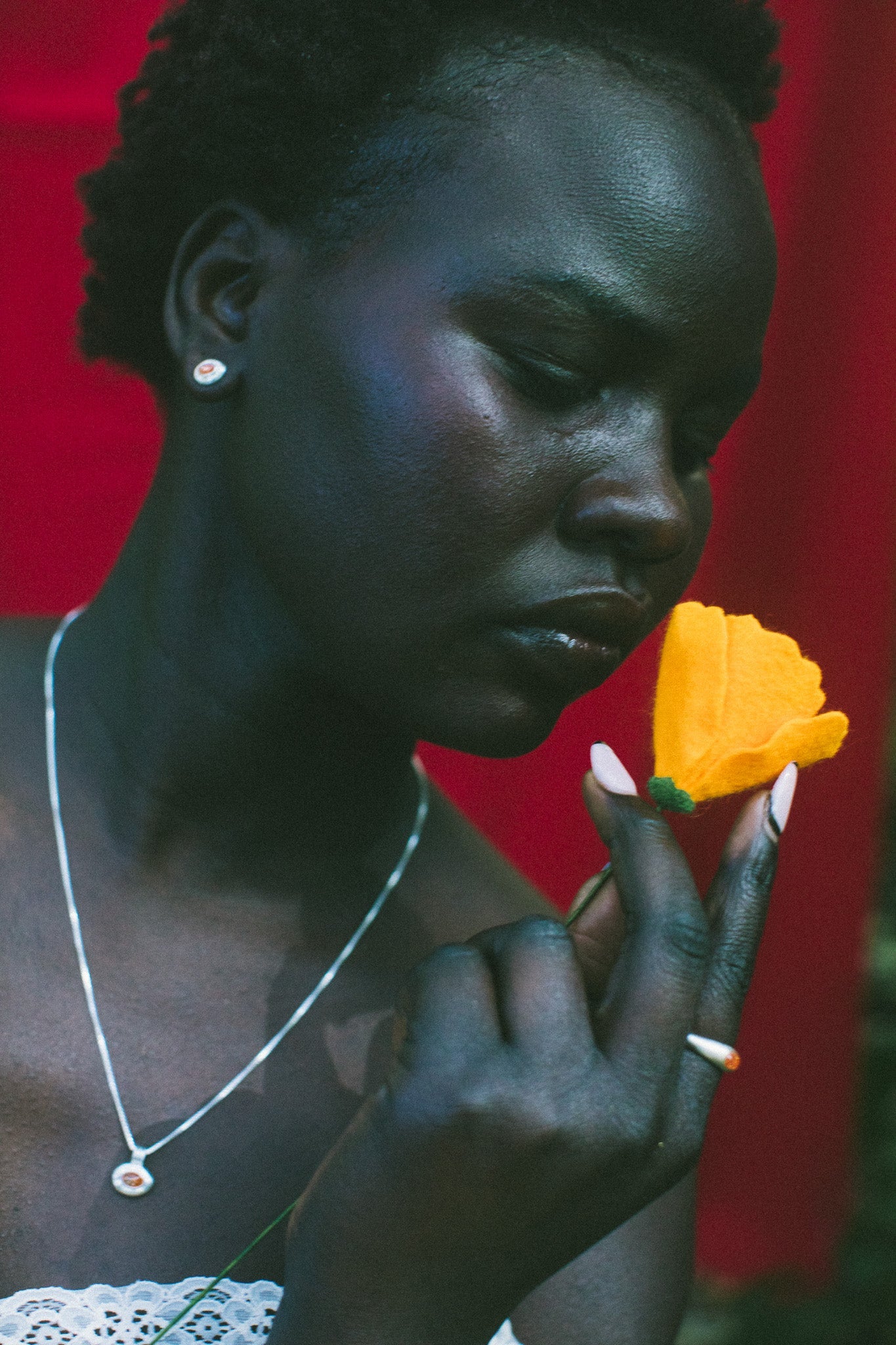 A close-up portrait of a woman holding a bright yellow flower near her face, wearing a silver necklace and earring against a vivid red background.