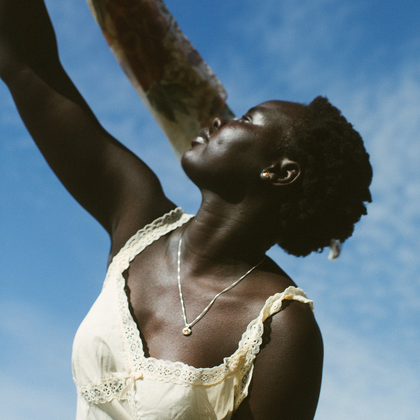 A woman wearing a cream lace-trim camisole top and a delicate gold necklace, looking upward against a bright blue sky.