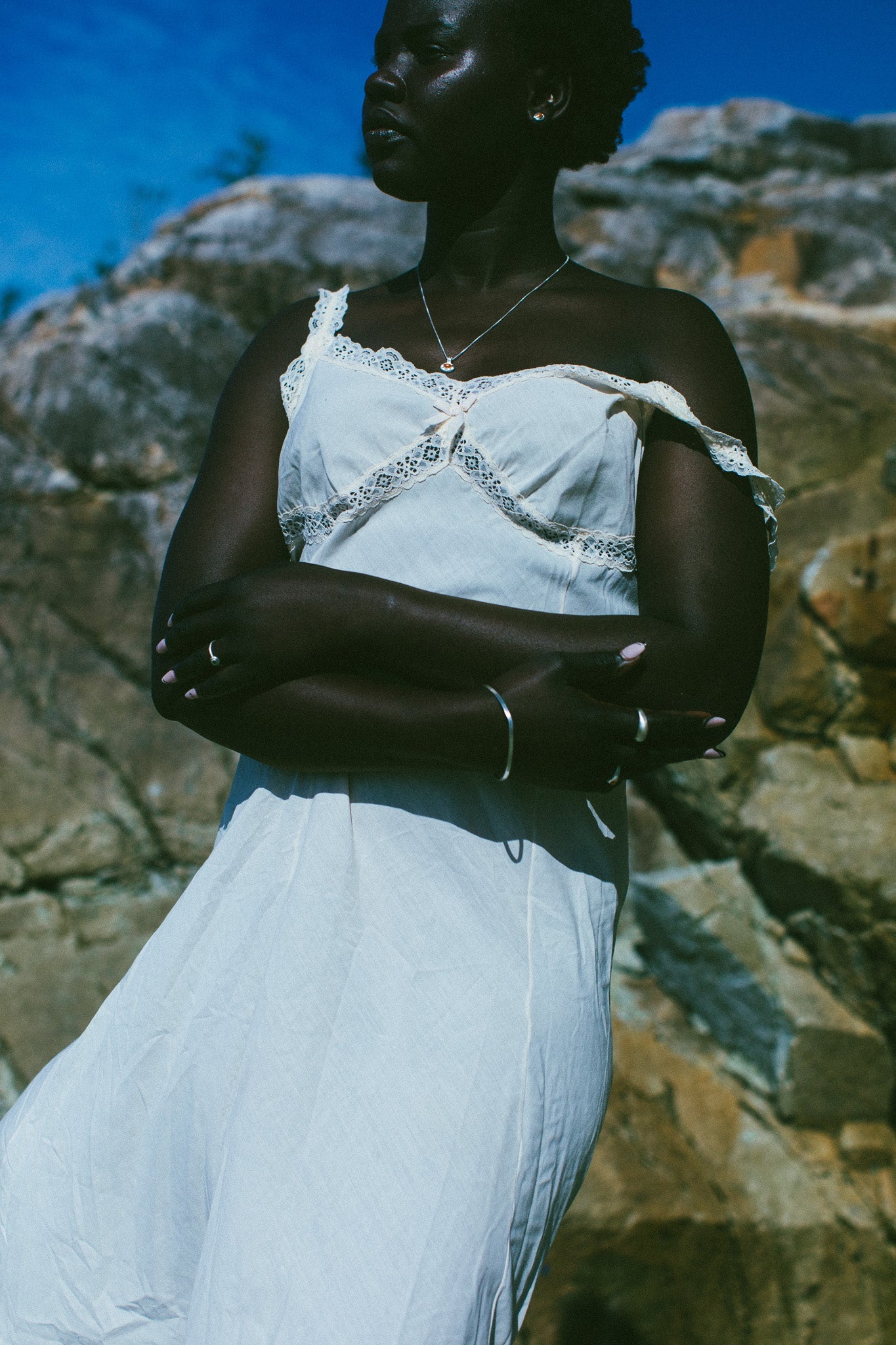 A woman wearing a white off-the-shoulder lace-trim dress stands outdoors against a rocky background under a blue sky.