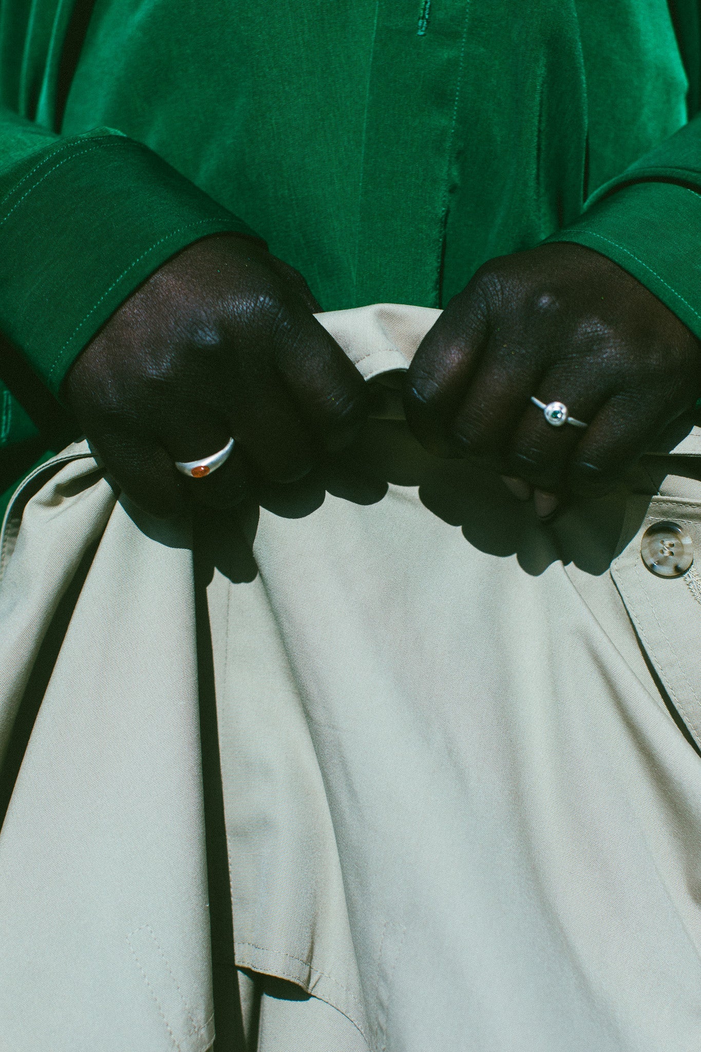 Close-up of a person's hands buttoning a light beige jacket or pants, wearing a vibrant green long-sleeve shirt and silver rings.