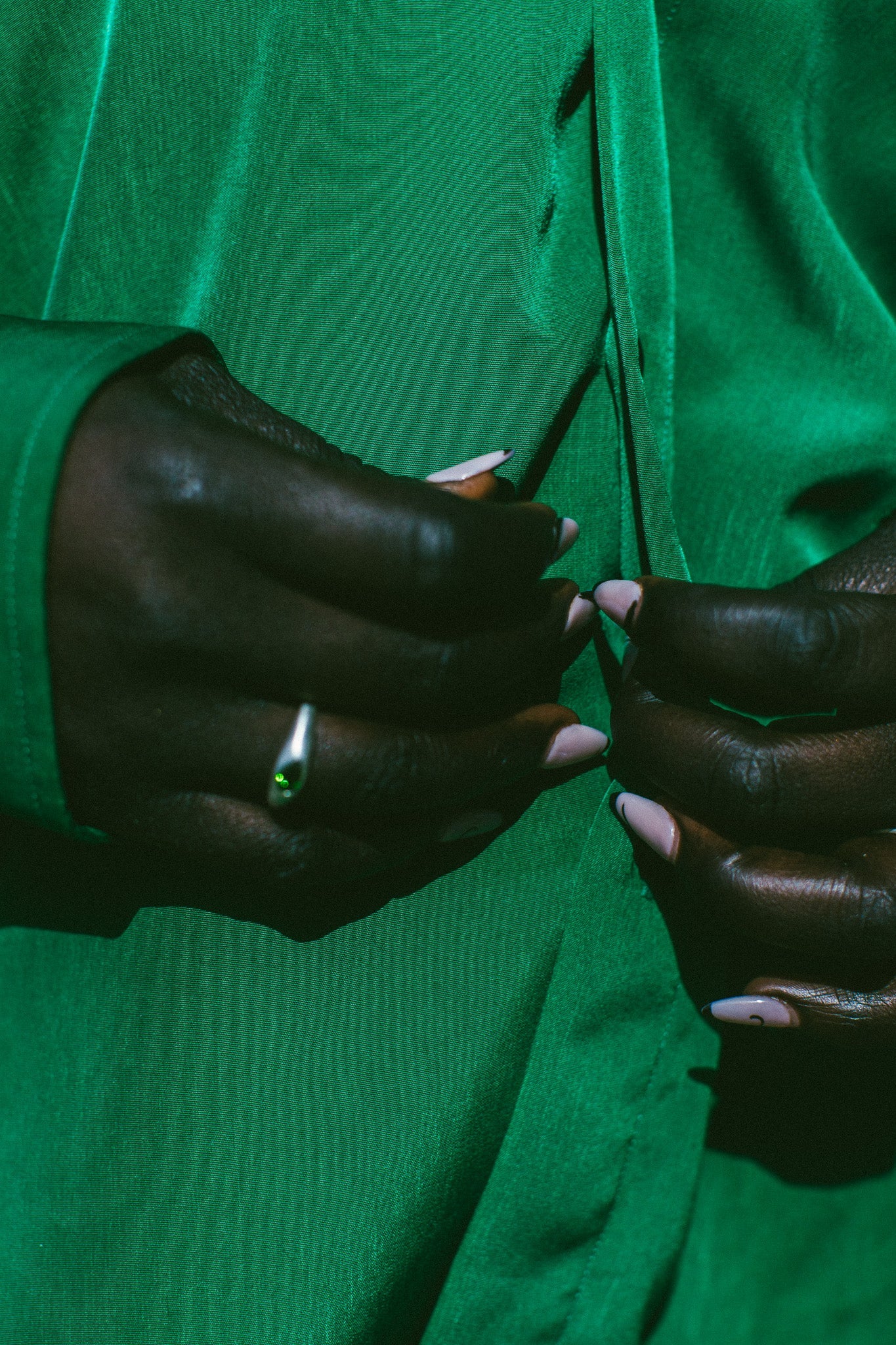 Close-up of a person wearing a vibrant green satin-like top or dress, with their hands buttoning the front. The person is wearing a silver ring with a green stone on one finger.