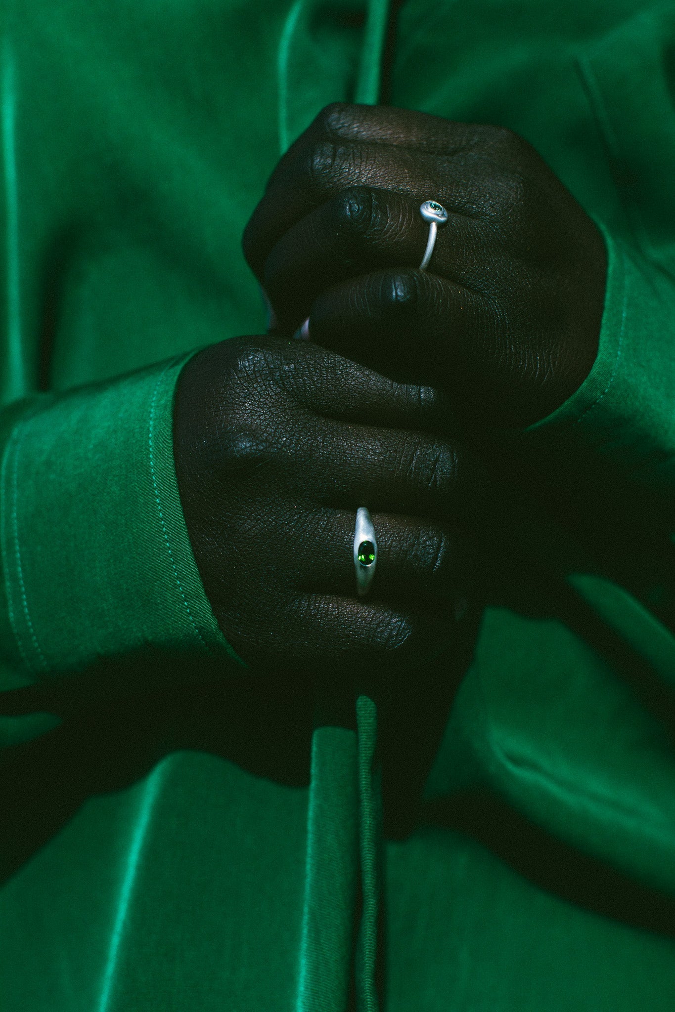 A close-up of a person's hands wearing two silver rings, one with a small round clear gemstone and the other with a small oval green gemstone, set against a dark green satin jacket.