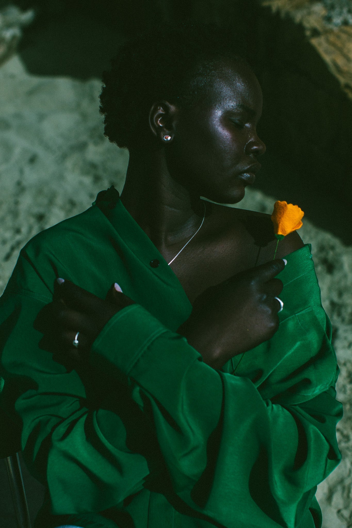 A woman in a dark green satin-like button-up shirt wearing silver earrings with an orange stone in the centre, 
holding an orange flower, set against a moody, shadowed stone background.