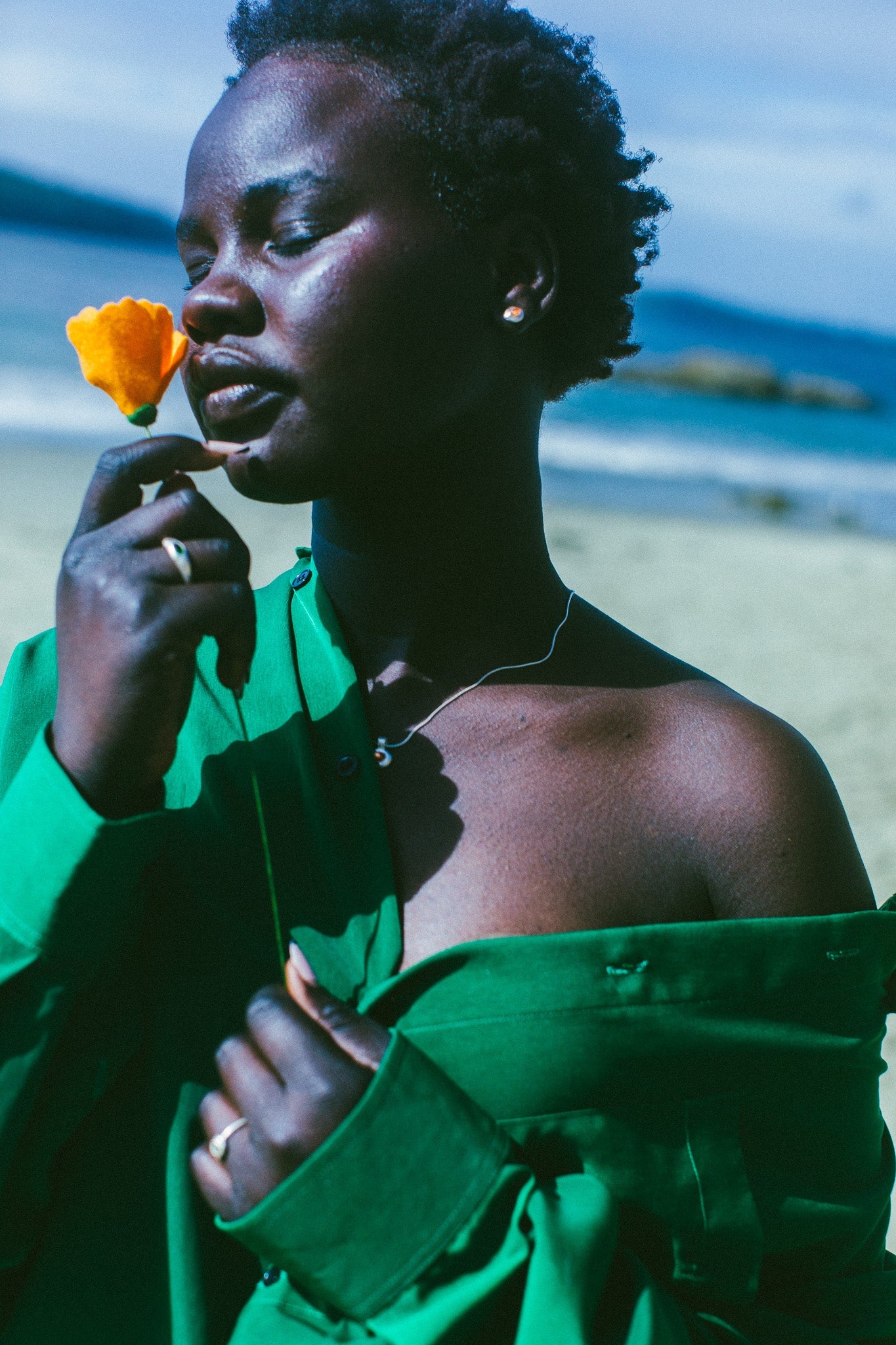 A woman on a beach wearing a bright green off-the-shoulder shirt, and silver earrings with an orange stone in the centre, holding a yellow flower near her face. The background shows sand and the ocean.