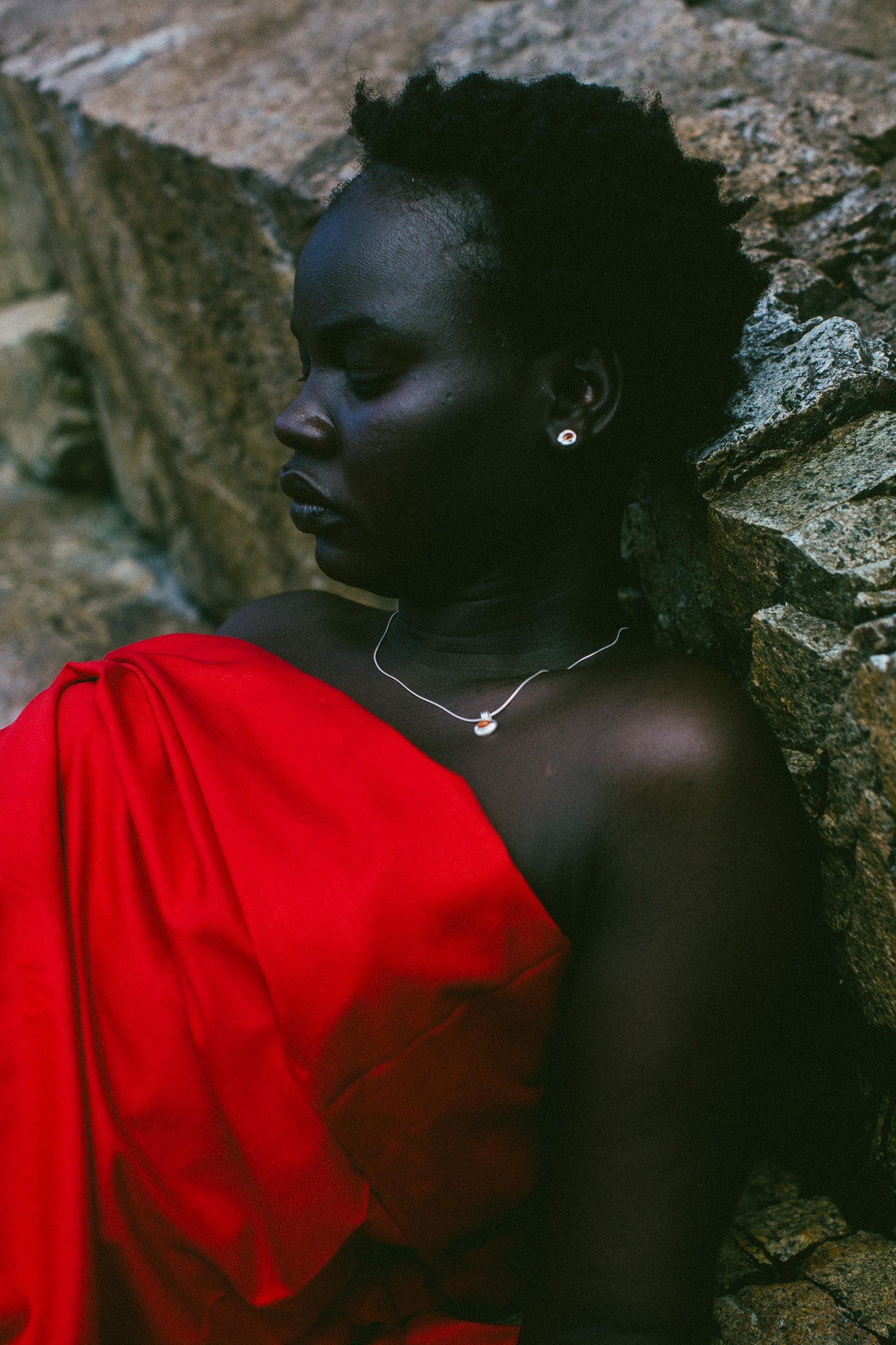A woman with short dark hair leans against a stone wall, wearing a bright red draped garment and a delicate silver necklace with a small oval pendant.