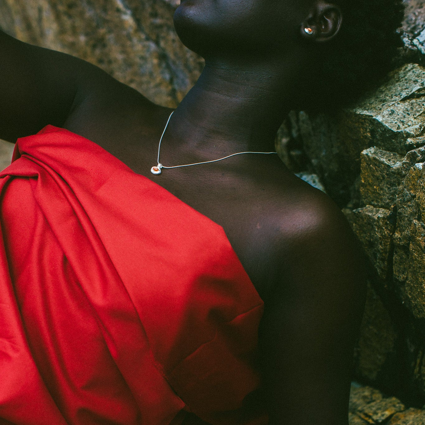 A close-up lifestyle shot of a model wearing a delicate silver pendant necklace and a matching silver stud earring, draped in a vibrant red garment against a dark, rocky background.