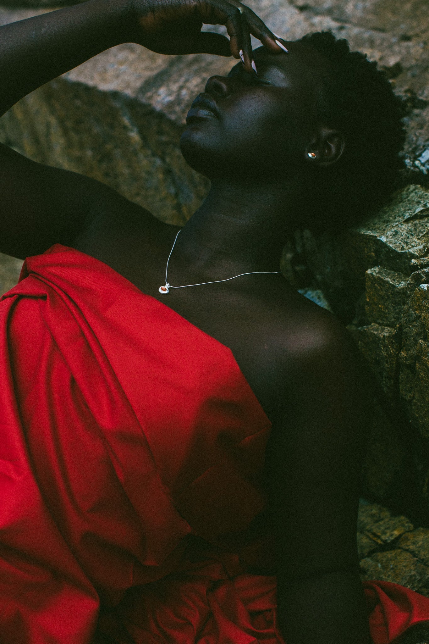 A woman reclining against a rocky surface, wearing a bright red draped garment and a delicate silver necklace.