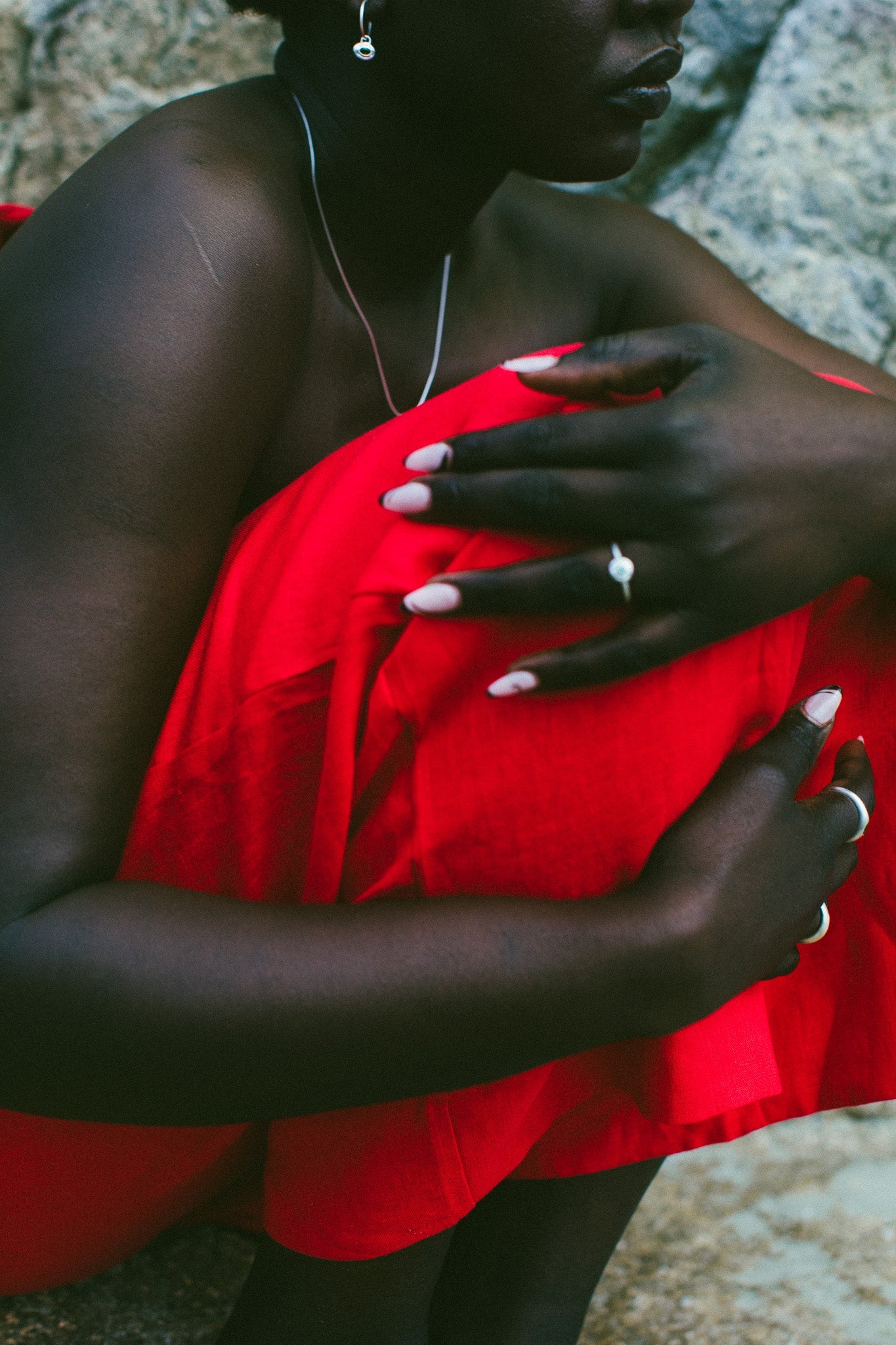 A close-up lifestyle shot of a model wearing a vibrant red strapless dress, sitting outdoors against a textured gray rock background.