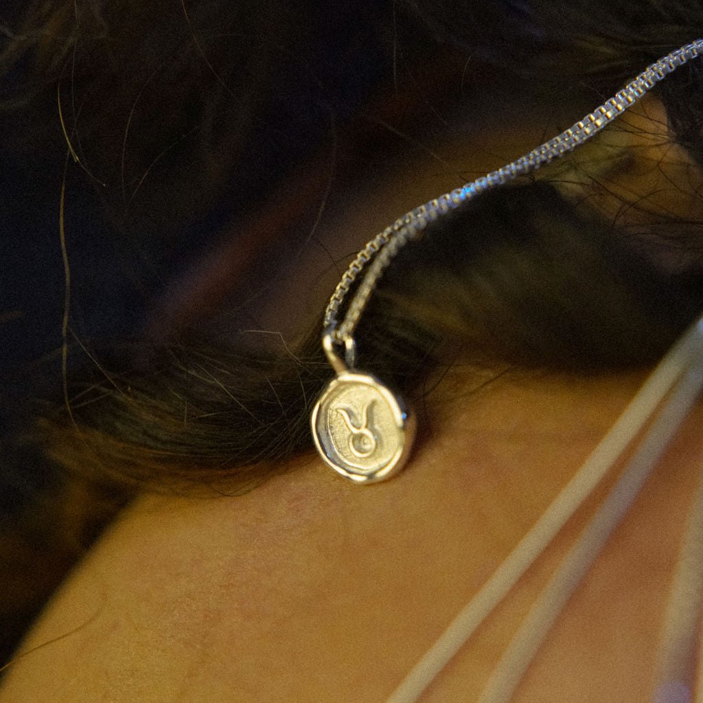 A silver necklace featuring a round pendant with a zodiac sign on it, attached to a delicate chain.