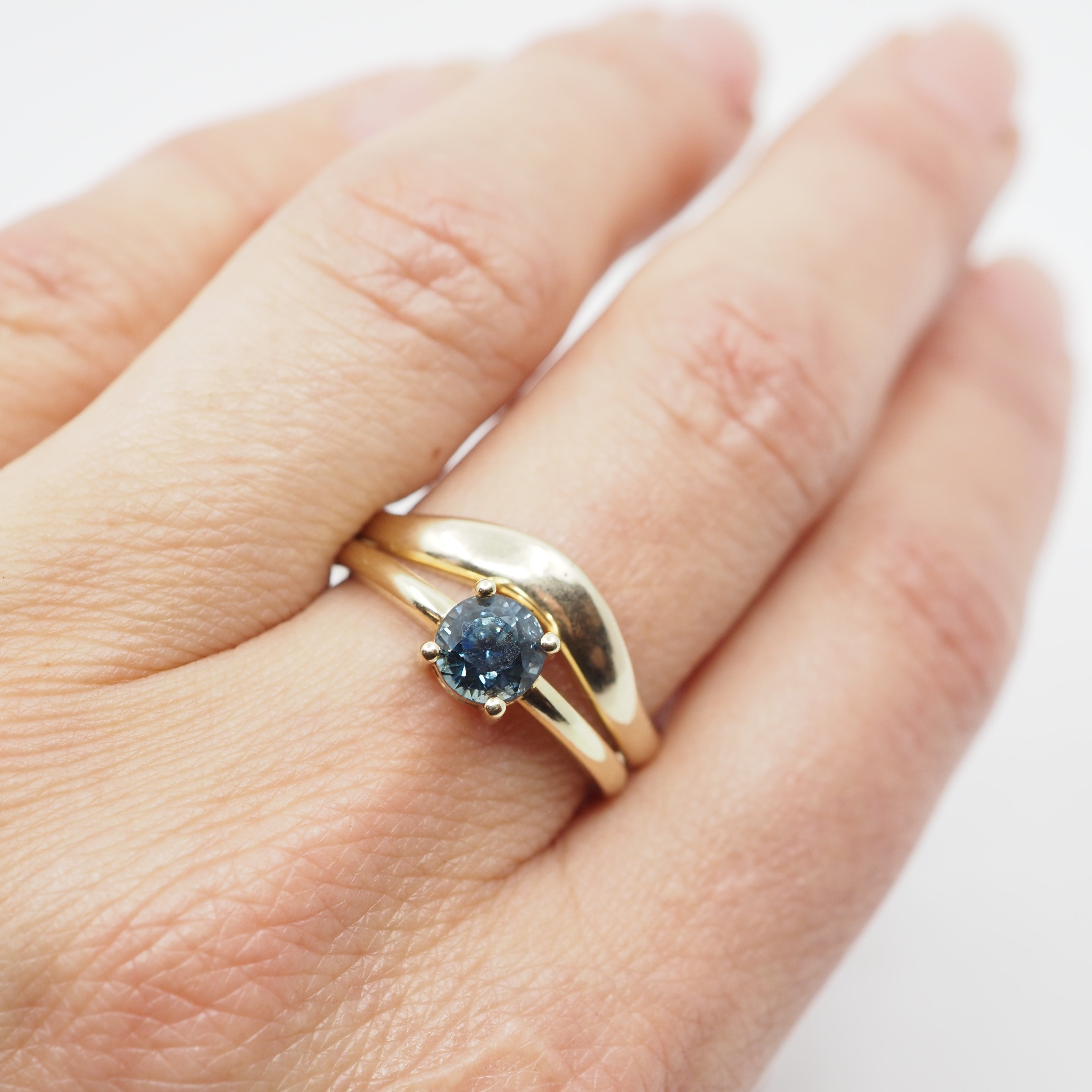 Gold ring stacked with a gold ring with a blue gemstone on a hand against a white background