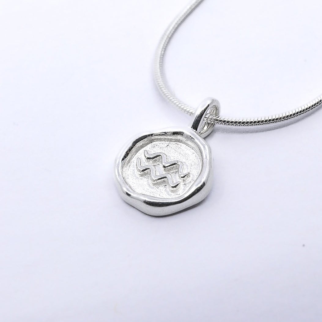 A silver-colored necklace with a round pendant featuring a zodiac sign symbol, attached to a chain.