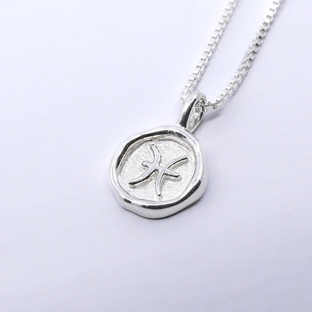 A silver-colored necklace with a pendant featuring a star sign emblem in a circular design, attached to a chain.