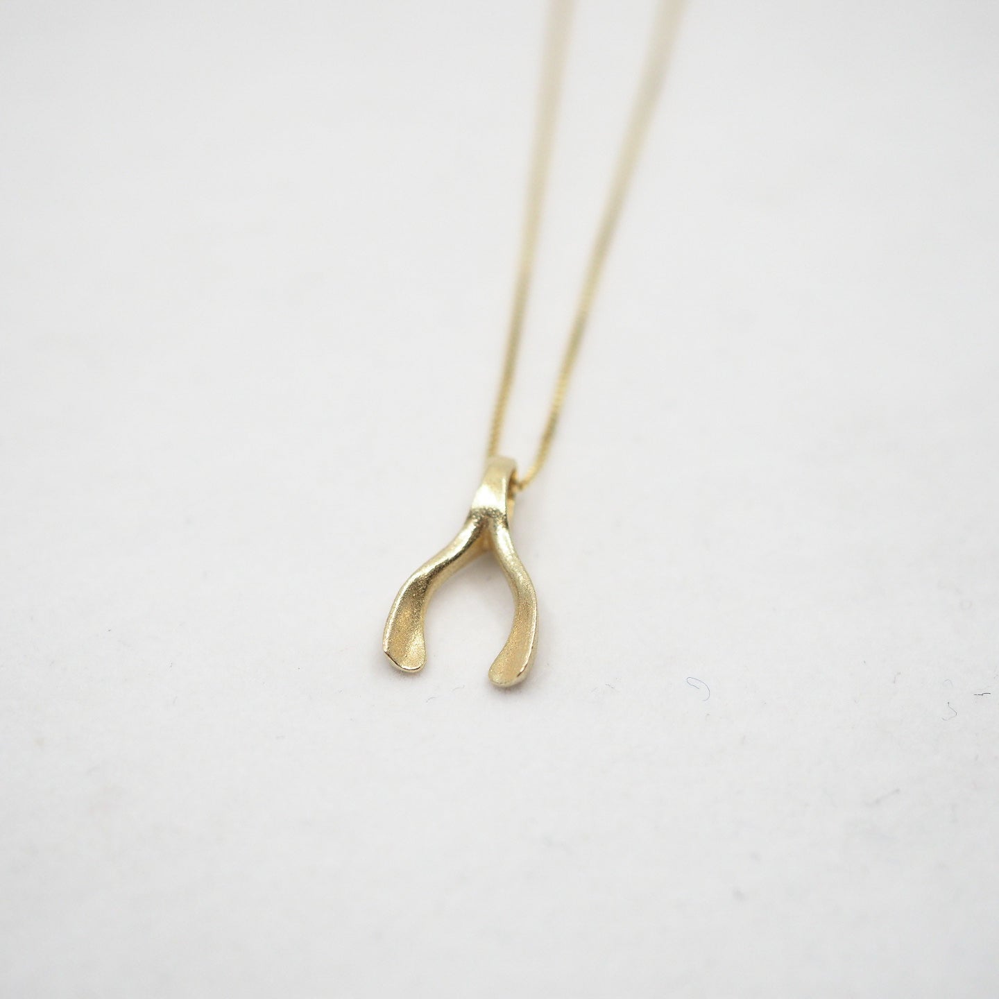 A delicate gold necklace with a small, wishbone pendant, photographed on a light gray textured background.