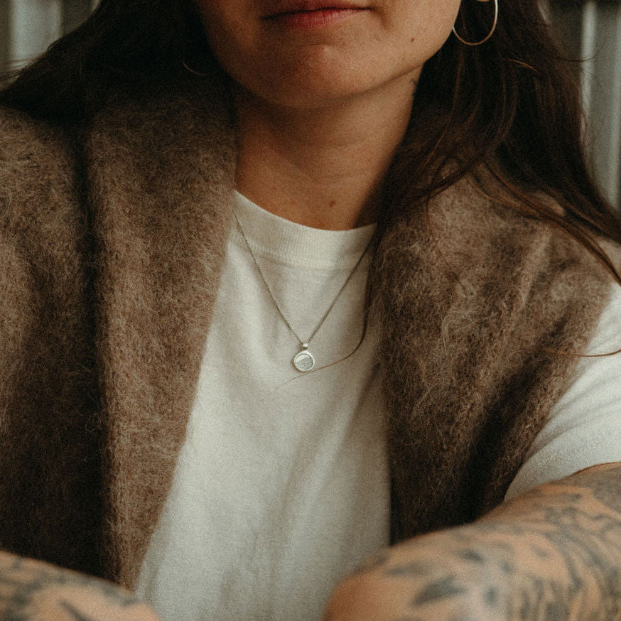 Person wearing a brown vest over a white shirt with tattoos on arms, sitting outdoors.