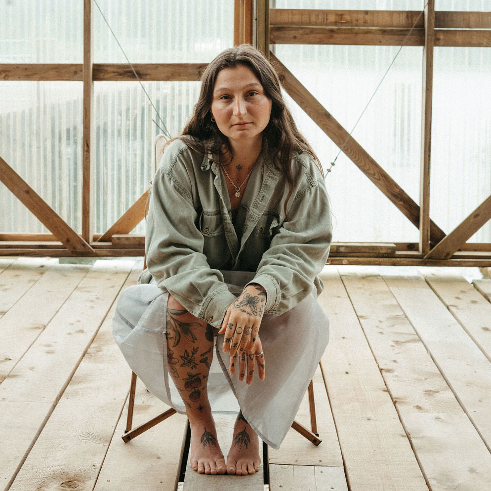 Woman sitting on a wooden chair in a rustic indoor setting with large windows, wearing the moyeha pendant. 